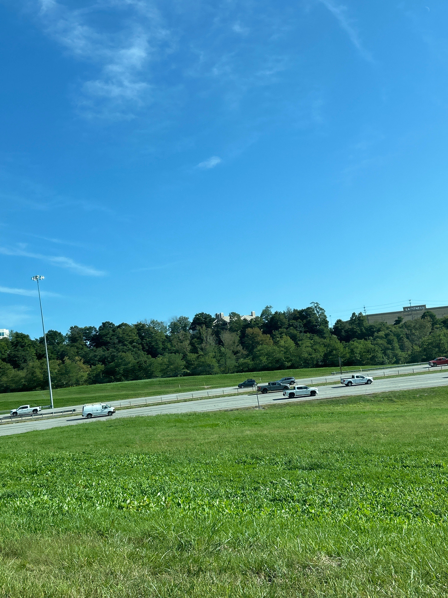 In Collier Township, Pennsylvania, a bright, sunny day is captured as seen from a grassy embankment overlooking a highway. The sky is a vibrant blue with wisps of white clouds. The highway, with multiple lanes, is active with vehicles, including white pickup trucks, a white van, and several cars. Beyond the highway, a dense line of green trees forms a backdrop, with some buildings and structures visible peeking through the foliage at the top of the rise. A tall street light stands prominently on the left side of the image. The foreground is dominated by a lush green grassy field.
