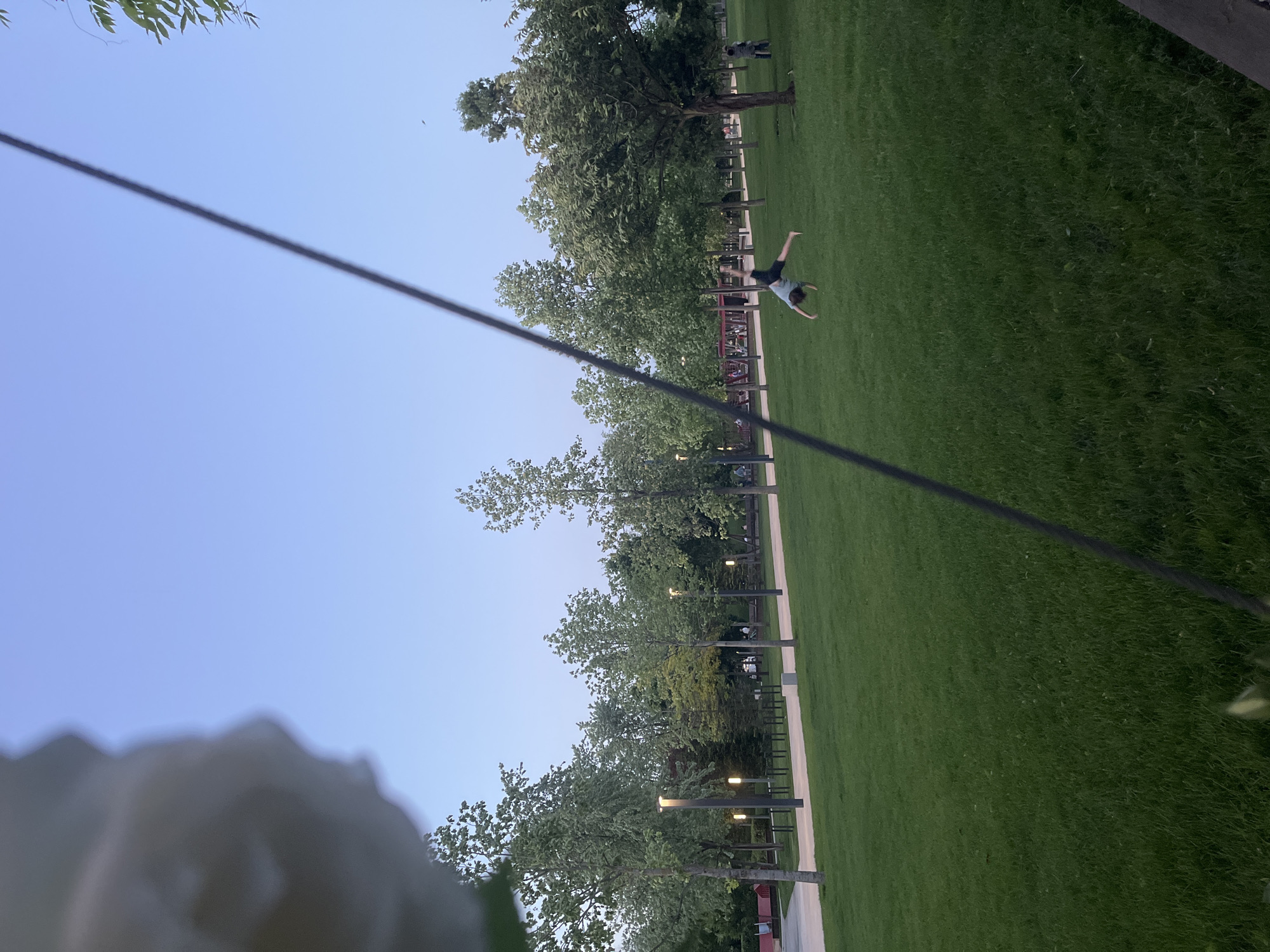 A park scene with green grass, trees, and a person doing a handstand is visible. The person is located in the middle ground of the image. There is a visible path with lamp posts along it. In the foreground, there is a partially visible object, possibly a statue. A power line cuts across the upper part of the frame. The sky is a clear, light blue. The scene is in Krasnodar, Russia.