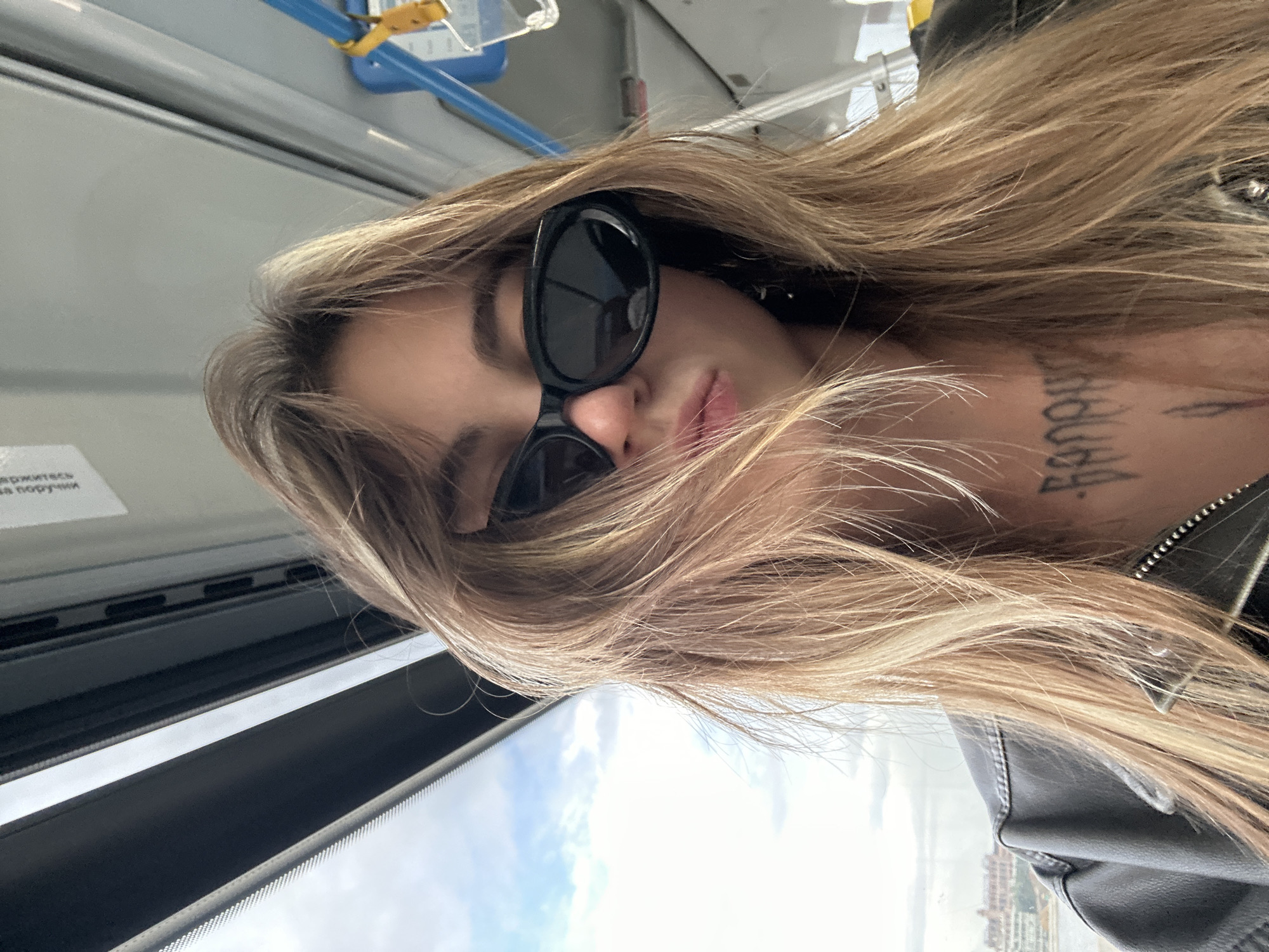 A young woman with long, blonde, wavy hair, wearing black sunglasses and a black leather jacket, is taking a selfie. Her hair is falling over her face, obscuring her eyes. She has a tattoo on her chest. She appears to be inside a vehicle, possibly a bus or train, with a window to her left showing a cloudy sky and a city skyline in the distance. The interior of the vehicle has blue and yellow grab bars. The overall impression is casual and candid. The city skyline suggests an urban environment, and the presence of public transport further supports this.