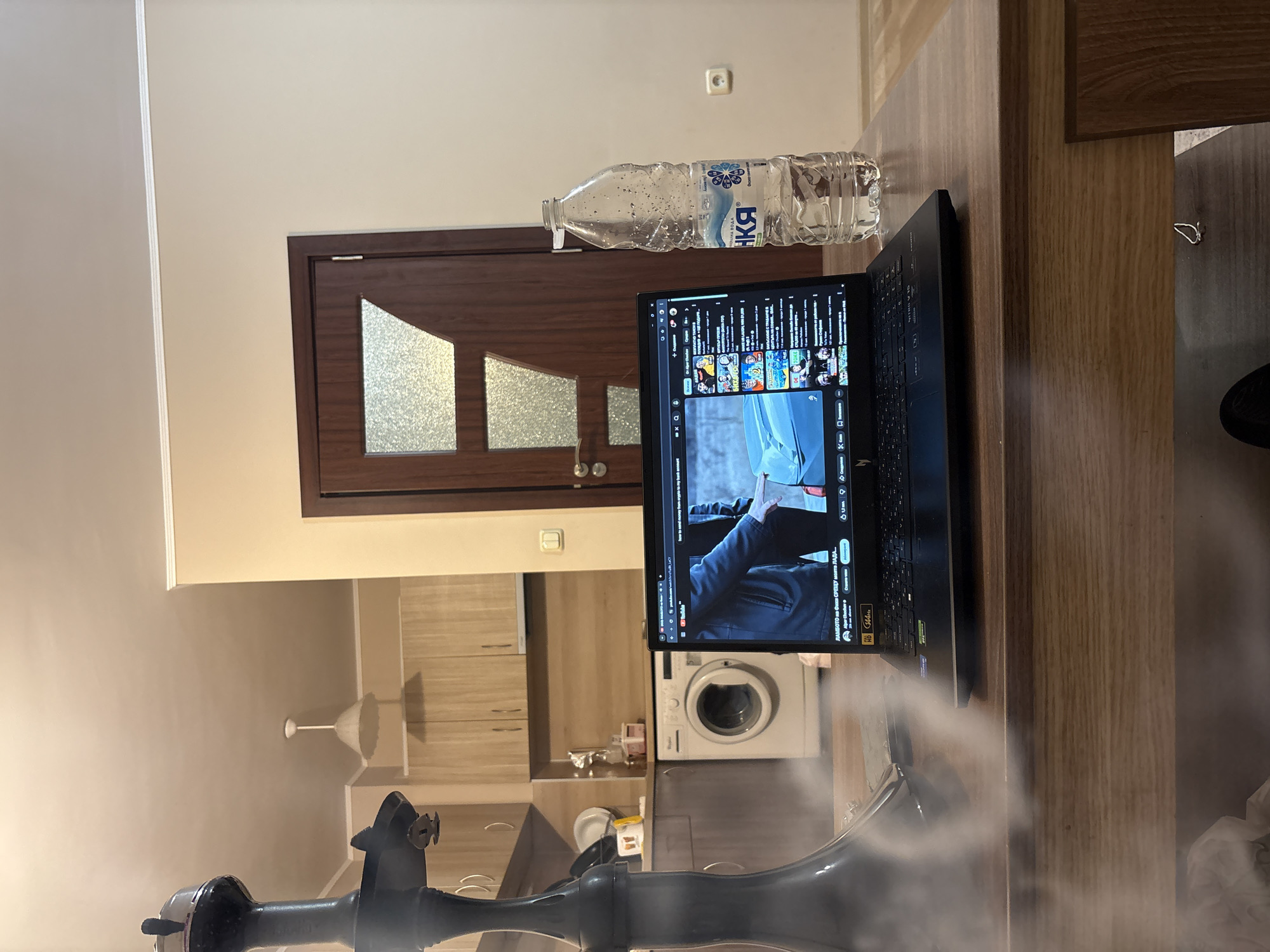 A still, indoor shot depicts a domestic scene. A laptop is open on a wooden surface, likely a table or counter, displaying a video on YouTube. To the left of the laptop is a clear plastic water bottle with a label, partially obscured. The scene unfolds in what appears to be a kitchen, as suggested by the washing machine and cabinetry in the background. A doorway with decorative glass panels is visible behind the laptop and water bottle. A hookah stands in the foreground, partially obscured by a cloud of smoke. The lighting in the room is soft, suggesting natural light. Based on the water bottle label and the Cyrillic text on the laptop screen, it is likely that the scene is located in Bulgaria. Given the popularity of hookahs and laptops globally, the image does not contain details specific enough to determine the exact location, even with knowledge of being in Bulgaria.