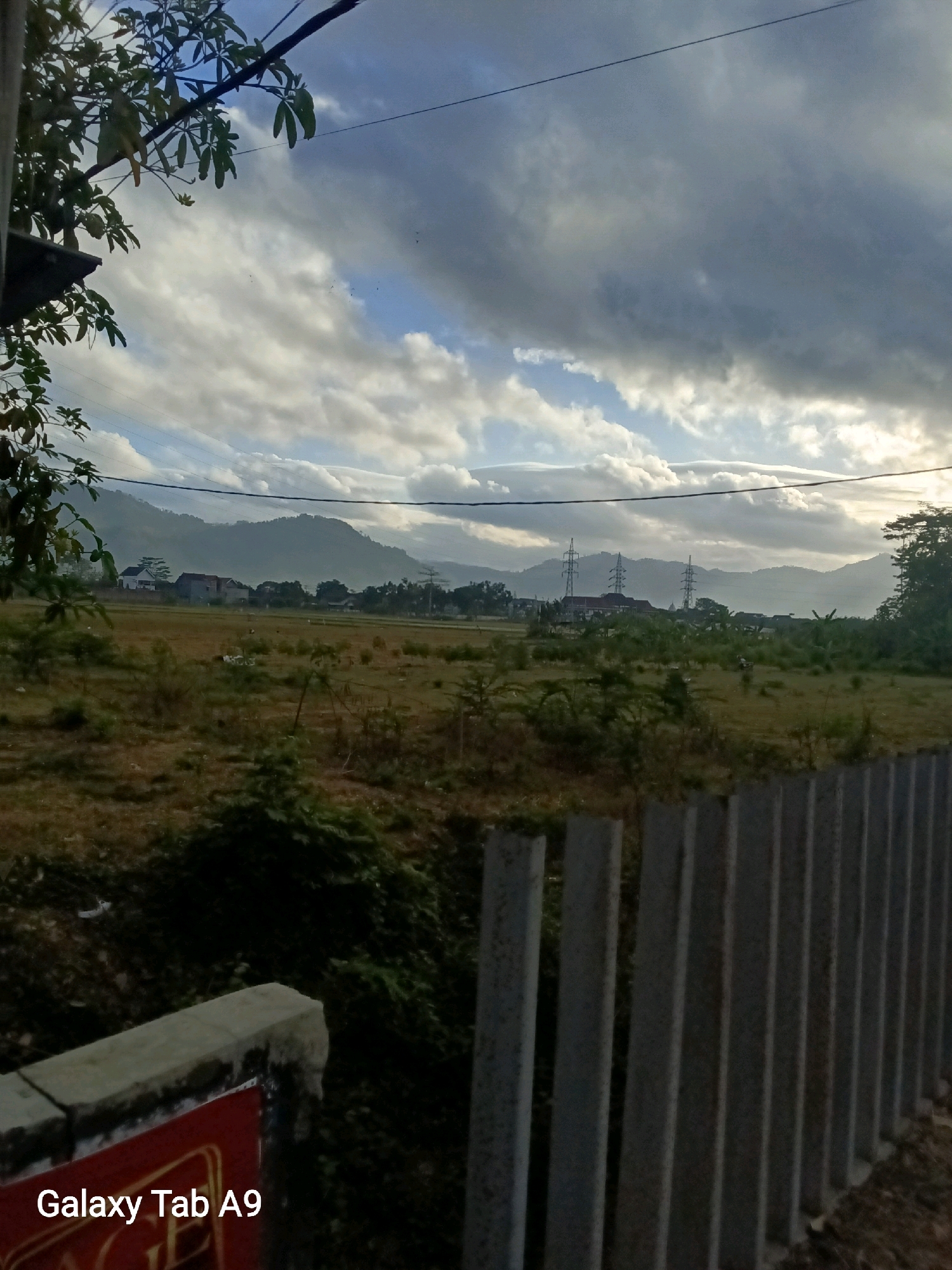 A wide landscape shot captures a serene rural scene in Trenggalek, Indonesia, under a dramatic cloudy sky. In the foreground, a concrete fence with vertical slats runs along the right side of the frame, with a partial view of a red sign bearing the text "Galaxy Tab A9" on the bottom left. Beyond the fence, a dry, grassy field stretches towards a cluster of houses and buildings in the middle ground, with tall transmission towers visible behind them. The background is dominated by rolling hills and mountains, softened by the atmospheric haze. Overhead, thick, grey clouds with patches of blue sky create an impression of impending or recent rain. The overall lighting suggests either early morning or late afternoon. No people are visible in the image.
