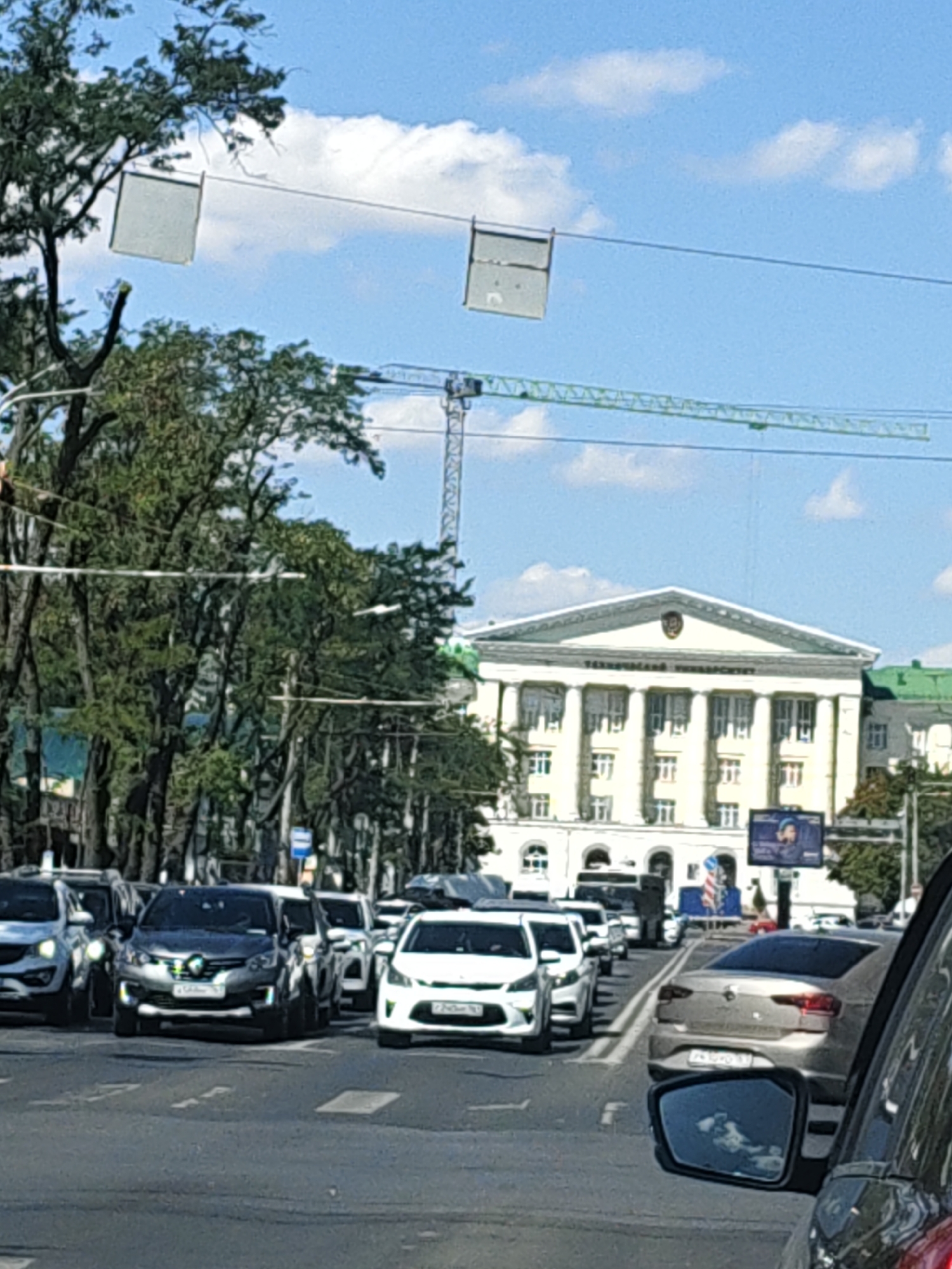 The image shows a street scene in Rostov-on-Don, Russia, on a sunny day with a blue sky and scattered clouds. In the foreground, several cars are stopped at a traffic light or in congestion on a paved road. The cars are of various makes and models, including a dark-colored sedan in the center, a white SUV to its left, and a light-colored sedan on the right. The perspective is from inside a vehicle, with a side mirror reflecting the sky and clouds.
In the mid-ground, a large, classical-style building with columns and a pediment is visible. This building appears to be a prominent landmark, possibly a university or administrative building, with the inscription "ТЕХНИЧЕСКИЙ УНИВЕРСИТЕТ" visible on its facade, identifying it as a Technical University. Trees line the street, partially obscuring the view of the building and other structures. A construction crane is also visible in the distance, suggesting ongoing development or renovation in the city.
In the background, there are more trees, buildings, and street infrastructure, including traffic lights and signs. The overall impression is of a typical urban environment with vehicular traffic and significant architecture.
