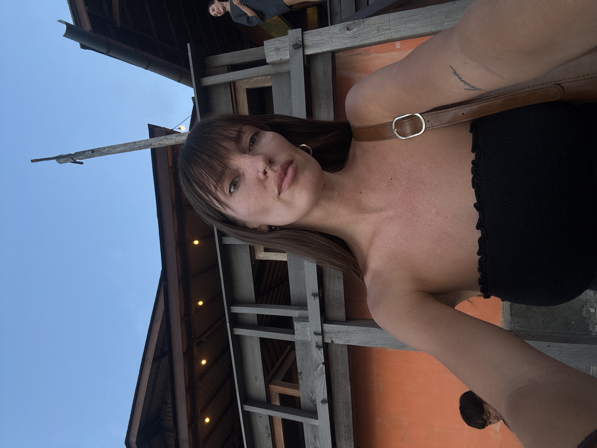 A low-angle selfie shot depicts a woman with dark hair and bangs, wearing a black strapless top and a brown leather strap. She has a nose ring and is wearing earrings. Behind her, an outdoor wooden structure with a red-orange wall and string lights is visible. Above the structure, a blue sky is present. In the background, another person can be seen on an upper level of the structure. The setting appears to be in North Kuta, Indonesia.