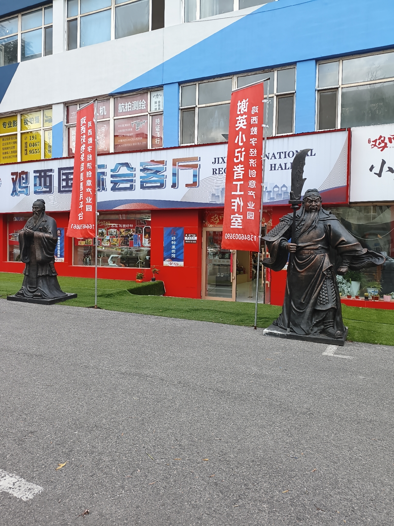 The image depicts an outdoor scene in the city of Jixi, China, in front of a building with a blue and white facade. Two large bronze statues of historical figures stand on a paved area with a grassy section in front of the building. The statue on the left is of Confucius, identifiable by his traditional robes and beard. The statue on the right is a warrior figure, possibly Guan Yu, characterized by his armor, long beard, and a weapon held aloft. Red banners with Chinese text are displayed in front of the building. The building itself features large windows and a red lower section with storefronts. The overall impression is that of a public space or a commercial area that incorporates historical or cultural elements.