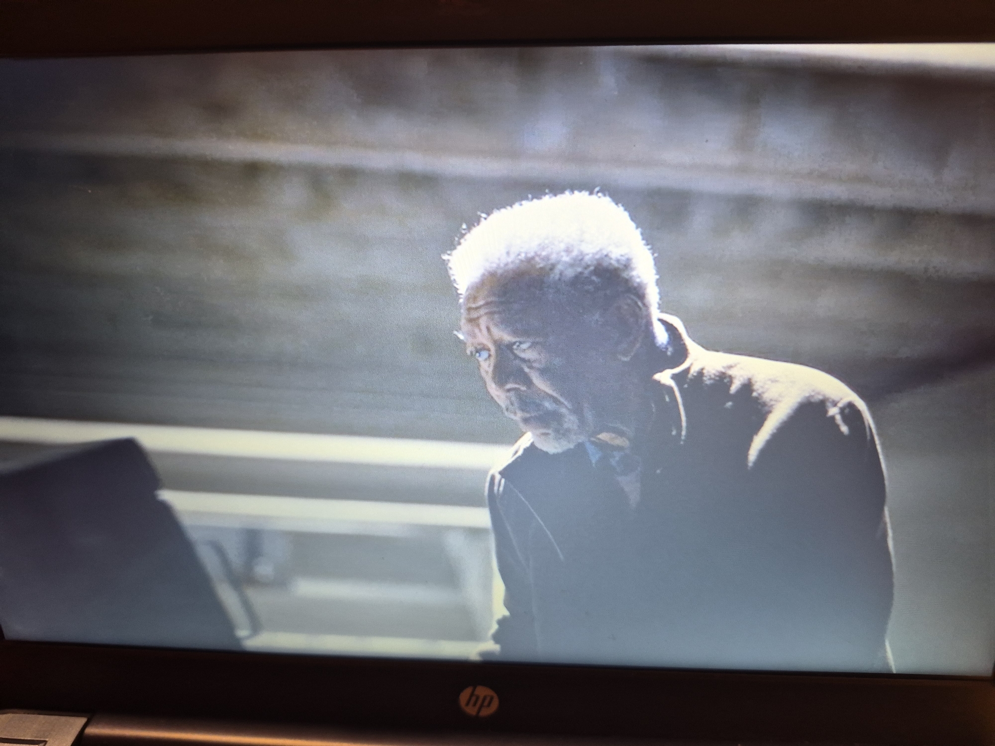 The image shows a man with white hair looking down and to the left. He is wearing a brown jacket over a blue shirt. The background is a dimly lit, industrial-looking space with horizontal lines that suggest shelving or beams. The lighting is dramatic, with the man's head brightly illuminated from above. There is a dark object, possibly a screen or a stand, in the lower left corner of the frame. The overall impression is one of contemplation or perhaps weariness. The image is a still from a video, as indicated by the texture and the faint reflections on the screen. There is no discernible location in Sandwell, United Kingdom, provided within the image itself.