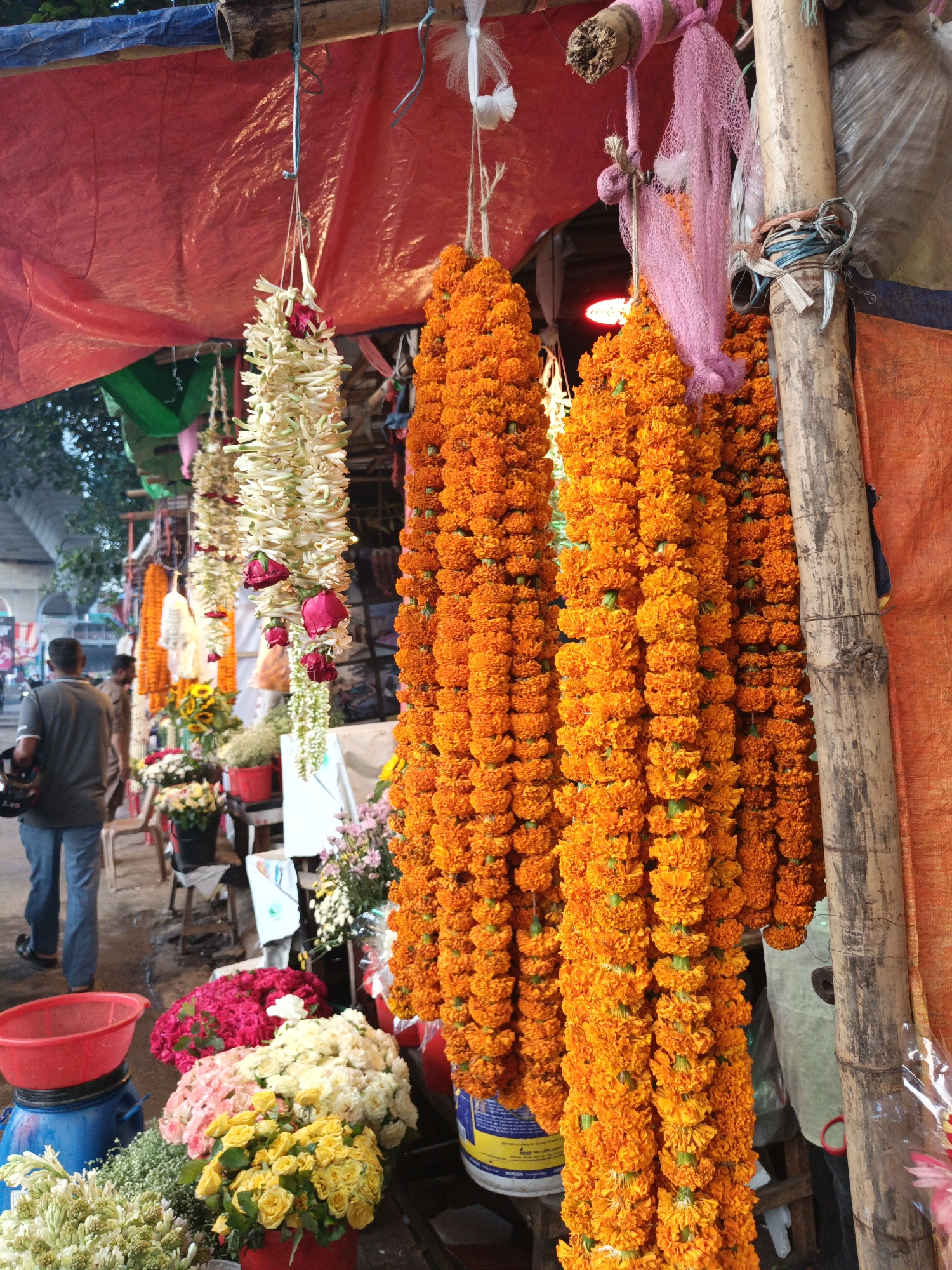 The image depicts a flower stall in Dhaka, Bangladesh. Hanging from a makeshift structure covered with a red tarp are garlands of orange marigolds and white flowers with red roses. Below, buckets and containers hold arrangements of various flowers, including yellow and white roses, pink carnations, and baby's breath. A man in a grey shirt and jeans walks away from the stall. The environment is bustling, suggesting a busy market or street scene.