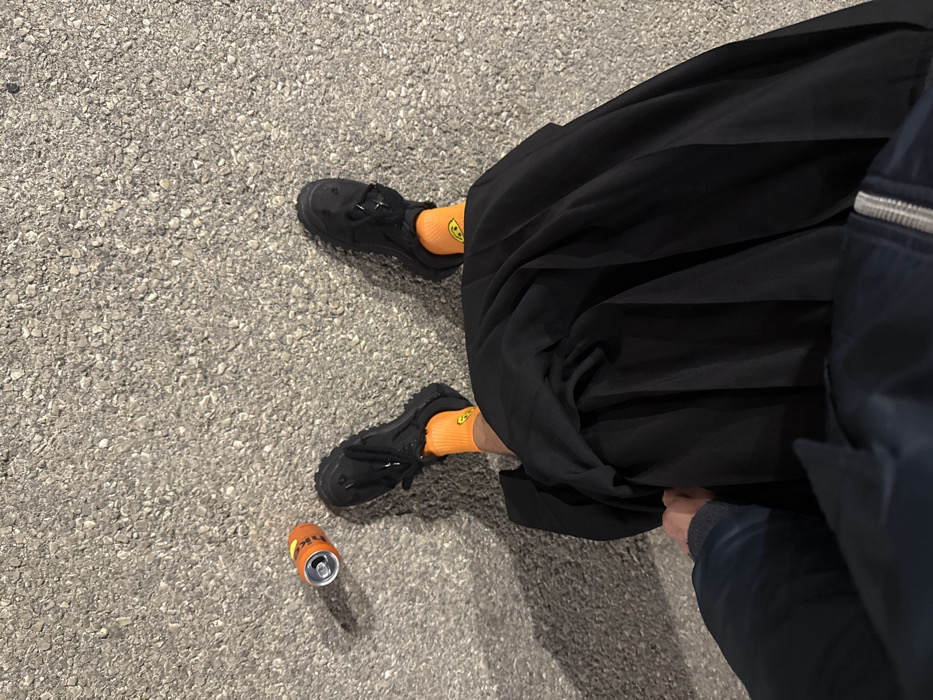 A person wearing black shoes and orange socks stands on asphalt near an open orange can. The person wears voluminous black pants or a skirt and a dark blue jacket. The ground is mottled gray. The image is taken from an overhead perspective.