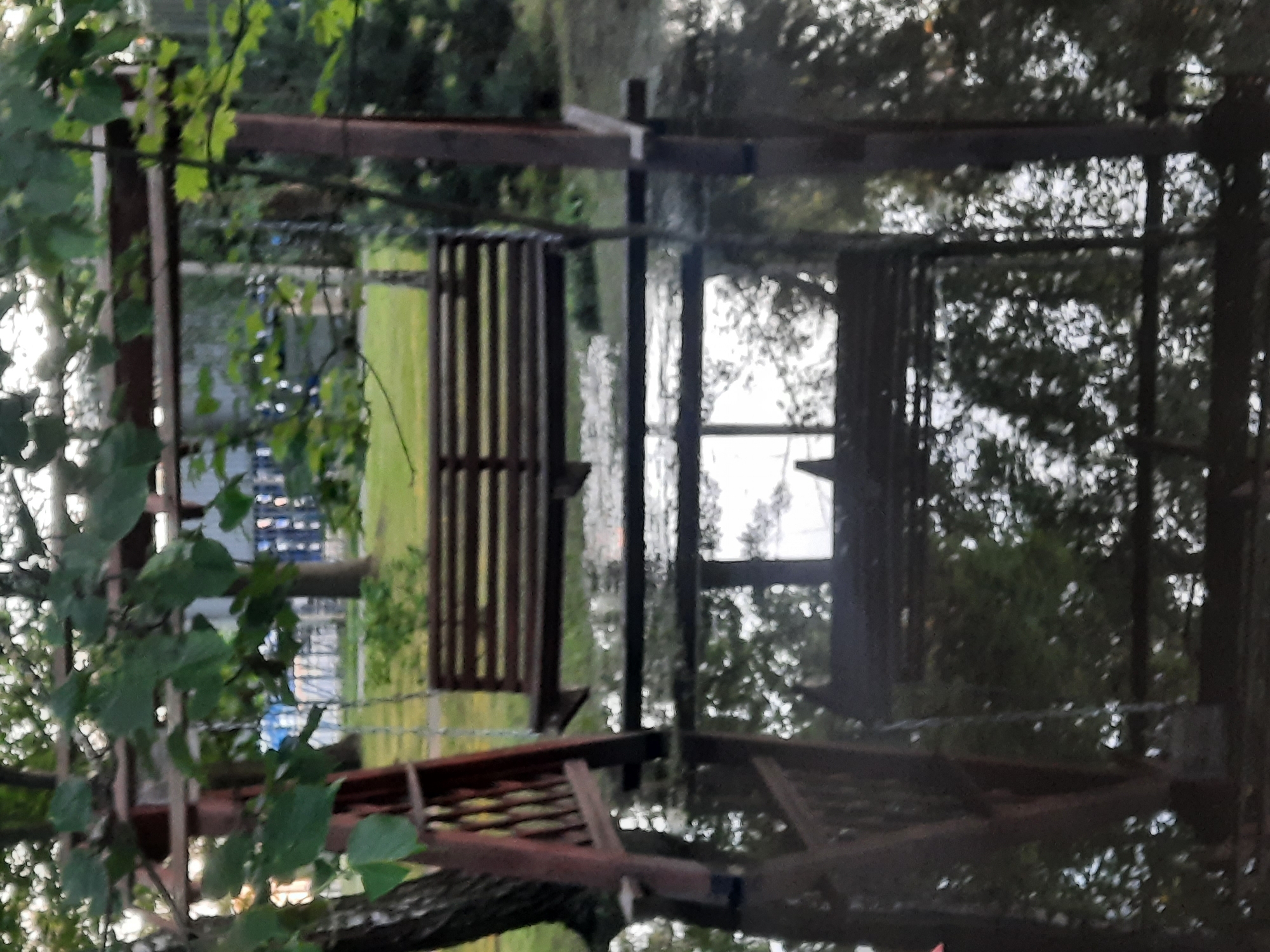 A wooden swing set with a wooden bench is partially submerged in water, suggesting a flood. The swing set appears to be outdoors, with trees and foliage surrounding it. The water reflects the swing set and the surrounding trees, creating a mirror-like effect. The scene is likely located in a park or backyard. The image is taken from a slightly elevated perspective, looking down at the swing set and the water. The lighting suggests it is either early morning or late evening, with soft, diffused light.