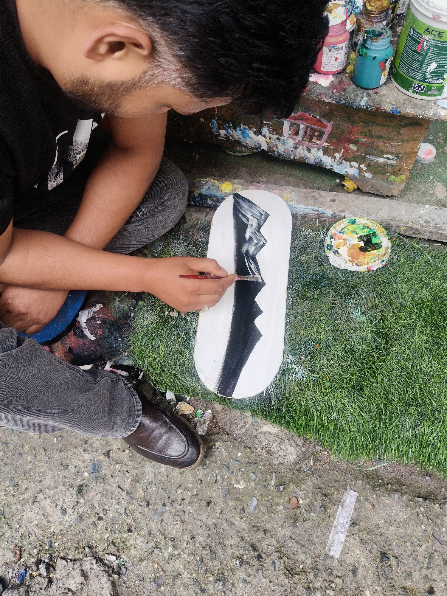 In Vashisht, India, a person is painting an abstract design on a white, oval-shaped object that appears to be a skateboard deck. The design features sharp, black, zigzagging lines against the white background. The painter, positioned on the left side of the frame, is crouched down, wearing a black t-shirt and grey jeans. Their right hand, holding a paintbrush with a red handle, is actively applying black paint to the deck. Nearby, on the green grass to the right of the deck, is a round paint palette covered in a mix of colorful paints. In the background, various paint bottles and containers are visible on a surface splattered with paint, suggesting an art-making environment. The ground beneath is a mix of concrete and dirt, with some scattered debris. The overall scene captures a moment of creative activity outdoors in Vashisht.