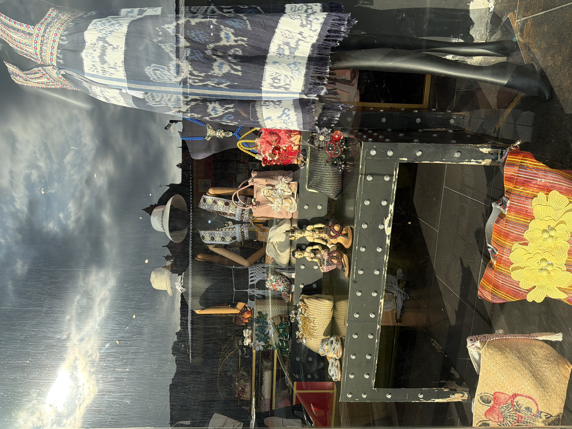 This media file captures a vibrant shop window display, seen through reflective glass that mirrors the sky and surrounding environment. The image, originally rotated 90 degrees clockwise, depicts merchandise commonly found in tourist areas, consistent with Kuta, Indonesia.
In the foreground, a dark mannequin wears a colorful dress or sarong made of patterned fabric, featuring intricate blue, white, and black ikat-style designs with some red and gold embroidered details near the top. Beside it, on a multi-tiered dark metal display unit studded with rivets, are various handcrafted accessories. These include a bright pink beaded purse, a black sequined clutch, several woven natural fiber bags (one with red embroidery, another with bright yellow floral appliques), and an intricately decorated belt. Two small wooden figurines, possibly traditional Balinese dancers, are also part of the display. Further into the window, several white woven hats are visible on stands, alongside other small decorative items and wooden mannequin hands.
Prominently reflected in the glass is a cloudy sky with a brilliant sun flare, indicating daytime. Silhouettes of distant buildings and possibly palm trees are also faintly visible in the reflections, suggesting an outdoor, street-side setting. No explicit text is visible. The overall scene conveys a cheerful, artisanal atmosphere.