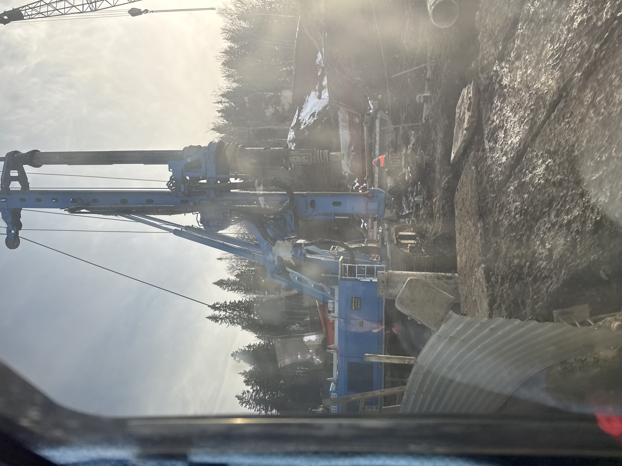 The image depicts a construction site in Perm, Russia, featuring heavy machinery including a large blue drilling rig. The rig is positioned on the edge of a dirt excavation. A crane is visible in the upper part of the frame. The background includes trees and hints of buildings through a slightly foggy or dusty atmosphere. Workers appear to be present near the rig. The photo seems to have been taken from inside a vehicle.