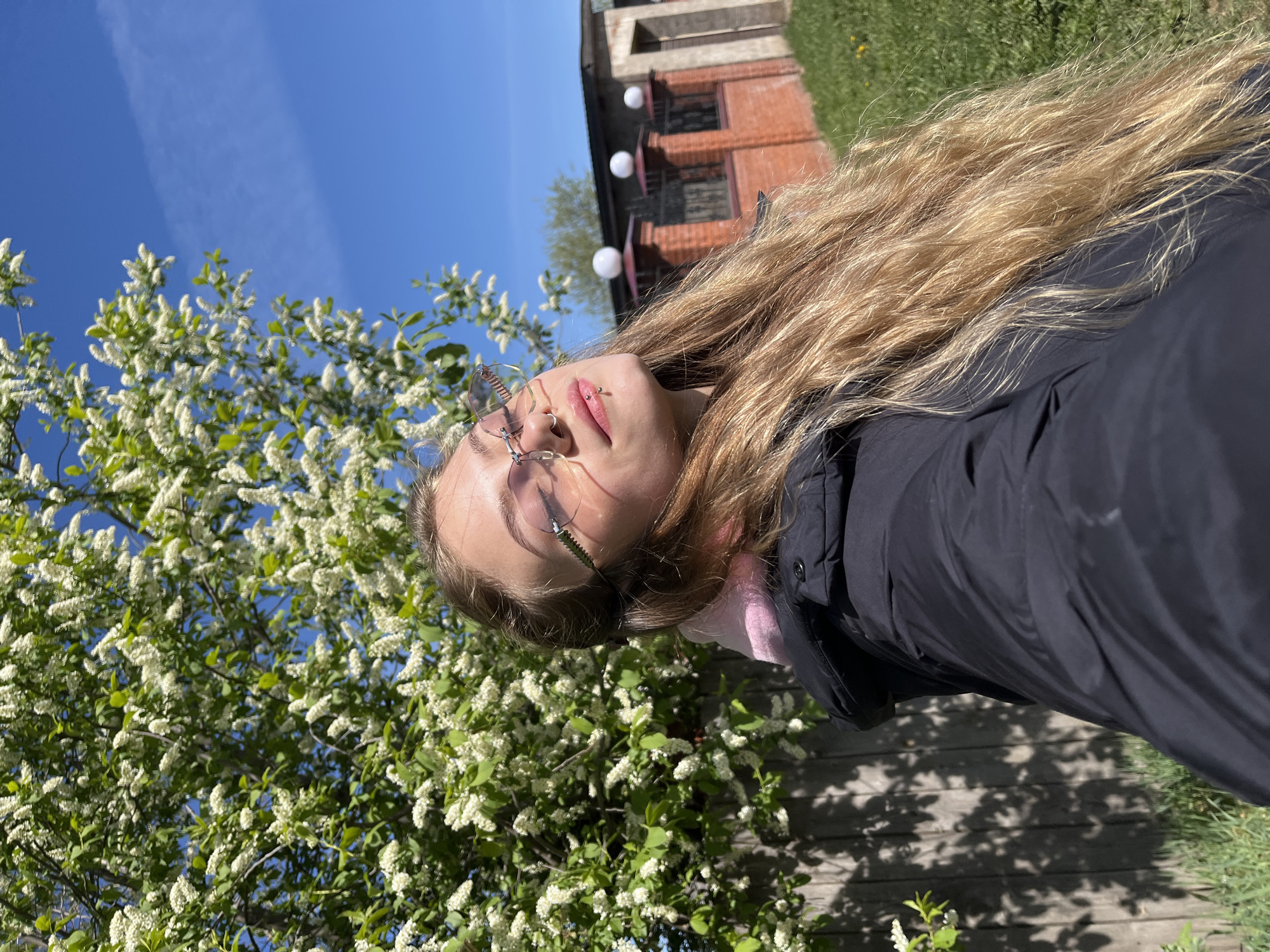 A slightly canted, medium-shot photograph depicts a young woman outdoors near flowering trees, potentially in the city of Фоки, Russia. The woman is wearing glasses, a black coat, and a pink shirt. She has long, blonde hair. Her face is angled upward towards a bright blue sky. A flowering tree with white blossoms is visible to the left of the woman. Behind her, a brick building can be seen, illuminated by a row of white, spherical lights. A wooden surface, possibly a fence or ground, with shadows cast by the trees is visible in the foreground. Green grass also appears in the upper right and lower right corners. The overall impression is a sunny, outdoor scene.