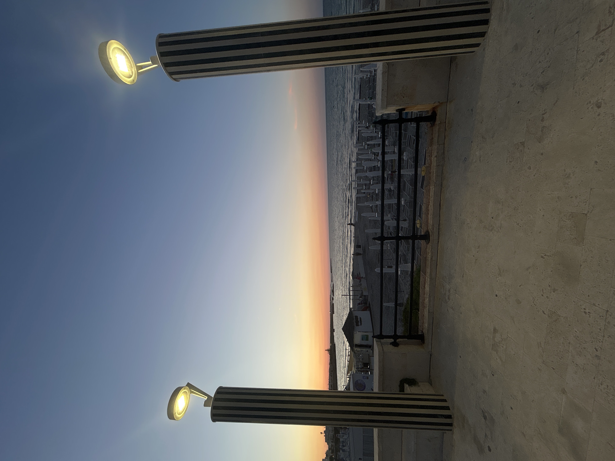 The image depicts a beach scene in San Foca, Italy, during sunset. Two prominent streetlights with black and white striped bases stand on either side of the foreground, framing a view of the Adriatic Sea. The sky exhibits a gradient of colors, transitioning from a deep blue at the top to shades of orange and pink near the horizon.
Below the sky, the sea appears calm with gentle waves. The beach is lined with numerous white beach umbrellas and chairs, some of which are closed, suggesting the end of the day. A small structure, possibly a beach bar or kiosk, is visible on the sand. A black metal railing separates the elevated promenade from the beach.
In the distance, a town or settlement is visible along the coastline, with some buildings illuminated. The ground in the foreground is paved with large, light-colored stone tiles. The overall atmosphere is serene and tranquil, characteristic of a coastal evening.