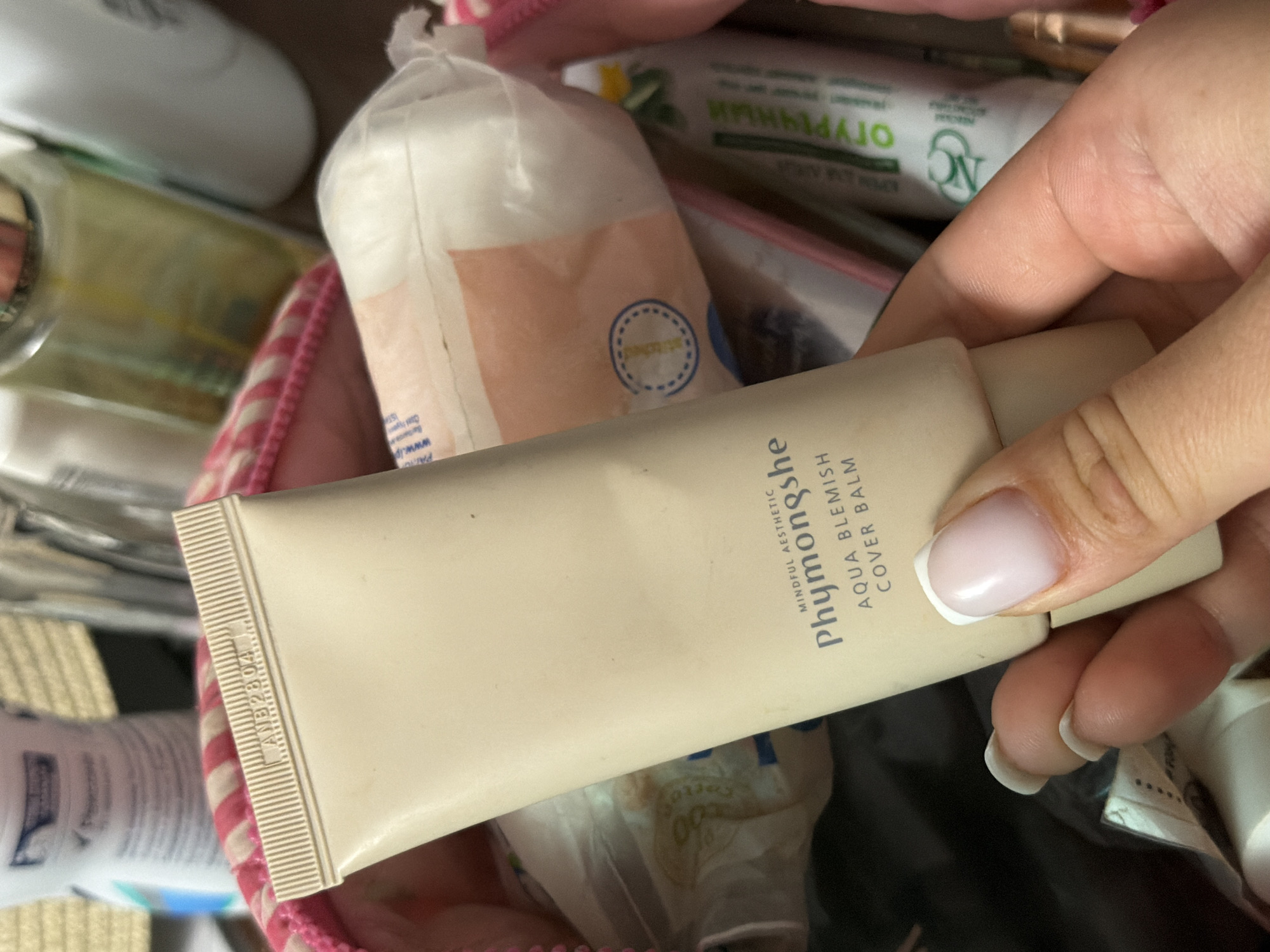 A hand with manicured nails holds a light beige tube of "Phymongshe Aqua Blemish Cover Balm". The tube has a ribbed cap and text indicating "MINDFUL AESTHETIC". In the background, a pink and white striped makeup bag is visible, containing various cosmetic items including cotton pads, a green and white tube with Cyrillic text, and other blurred products. The image appears to be taken indoors.
