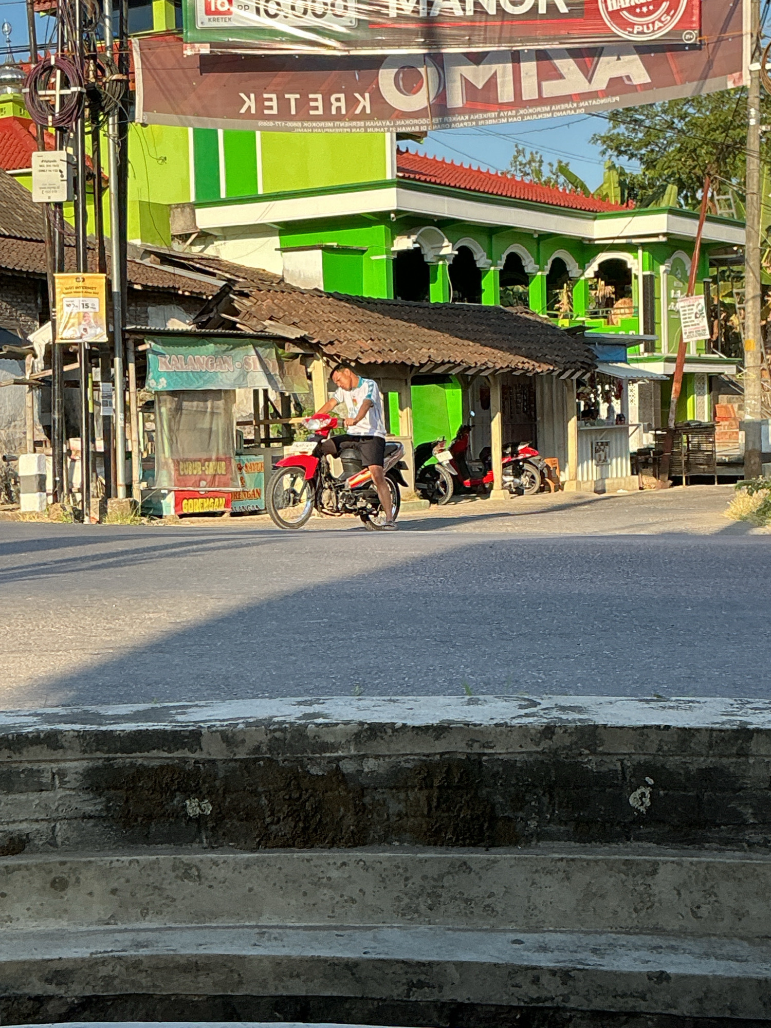 The image depicts a street scene in Kebonrejo, Indonesia, on a clear, sunny day. In the foreground, a paved road dominates the lower half of the frame, with a shadow of a tree falling across it from the left. Further back, a man is seated on a red motorcycle, wearing a white t-shirt and dark shorts. He appears to be looking towards the right side of the image.
Behind the man and motorcycle is a row of low-rise buildings with traditional tiled roofs. The building directly behind him has an open front with a counter displaying various items. A green sign with white text, possibly advertising "KALANGAN - SIRUP," is visible above this structure. Adjacent to this, to the right, another building with a similar roofline is seen, and further to the right, a taller, vibrant green building with white arched colonnades stands out. This green building has multiple stories and is decorated with white trim.
Above these buildings, a large banner is stretched across the scene. The banner is primarily white and red, with text that is partially obscured. Visible text includes "MANOR," "KRETEK," and some smaller text in Indonesian, which may indicate pricing or product information.
Several utility poles with wires are present on the left side of the image, one of which has small signs attached. Two other motorcycles are parked near the buildings on the right. The overall environment suggests a commercial or residential area within a town or city. The time of day appears to be late afternoon or early morning, given the low angle of the sunlight and the long shadows.