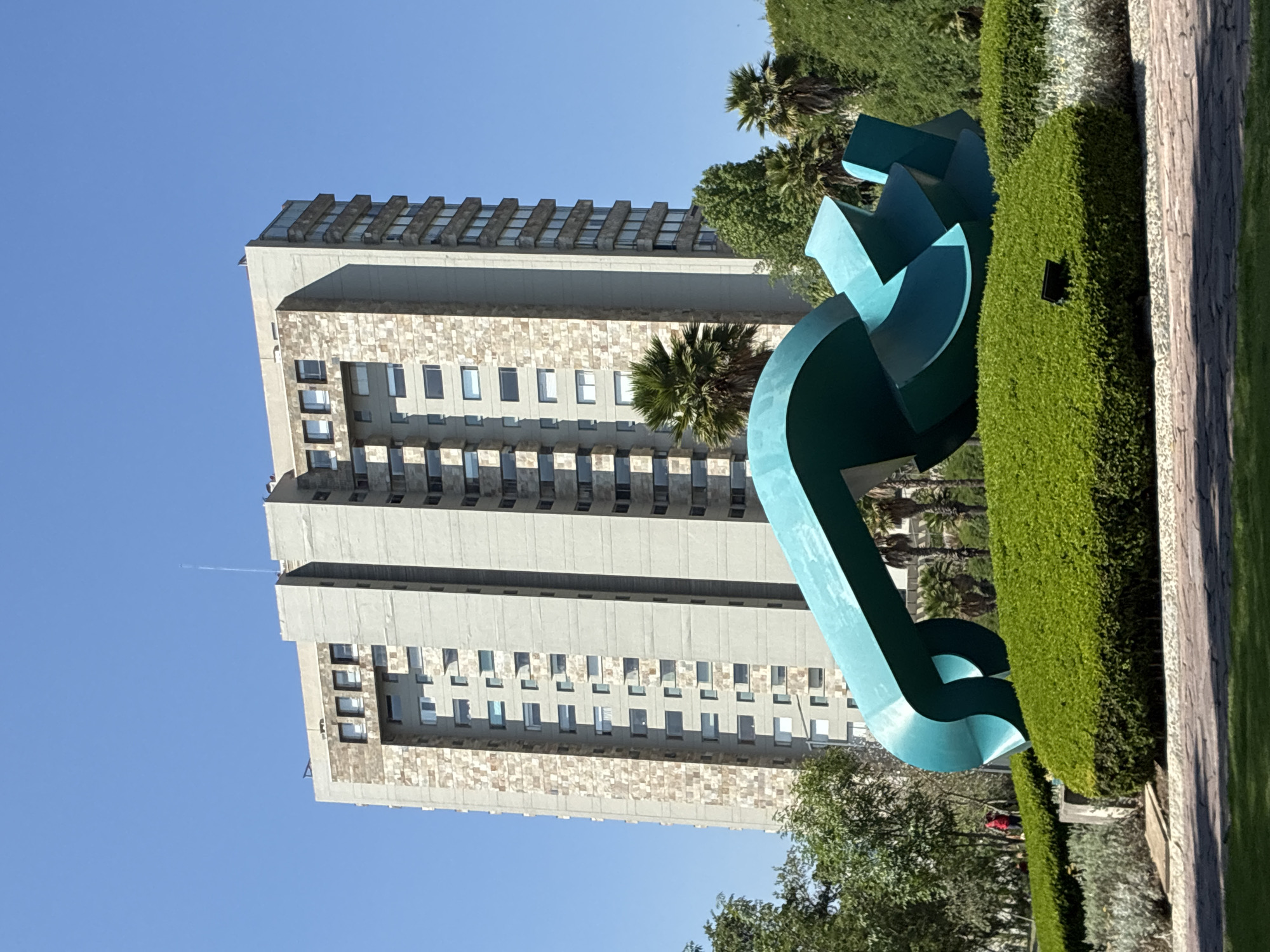 The image depicts a tall building in Huixquilucan de Degollado, Mexico, with a distinctive modernist architectural style, featuring a light-colored facade with textured stone accents and a repeating pattern of windows. In the foreground, a large, abstract sculpture painted in turquoise blue stands next to a manicured hedge. The sky is clear and bright blue, indicating a sunny day. The presence of palm trees and other greenery suggests a well-maintained urban landscape. A person wearing a red garment is partially visible in the background behind the sculpture and shrubs.