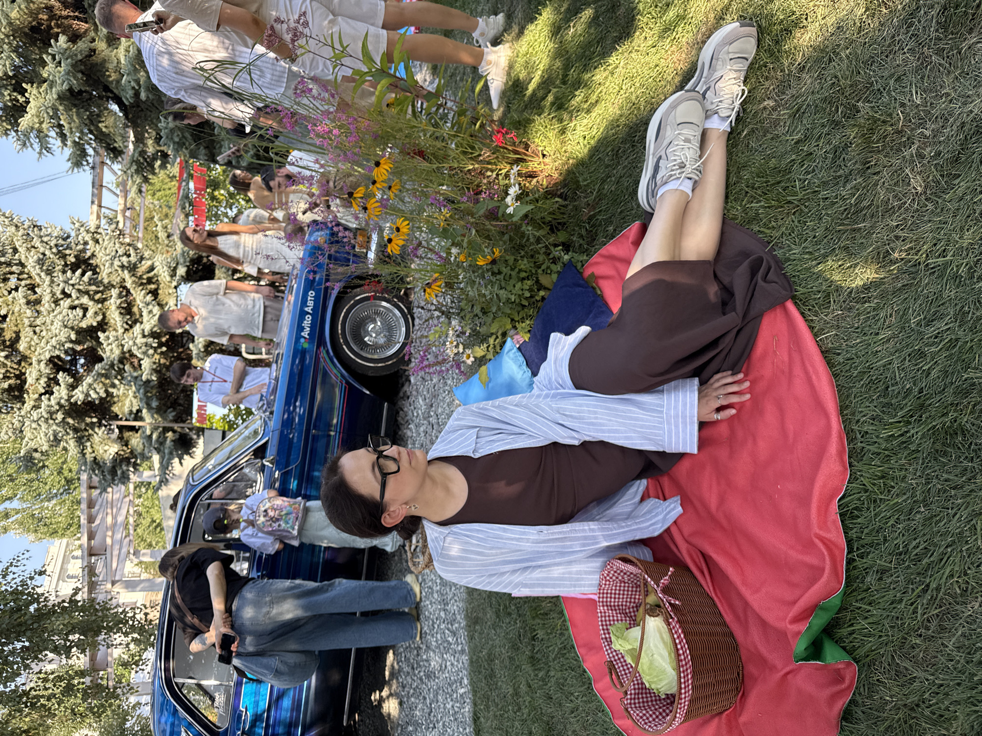 In Kazan, Russia, a woman with dark hair, glasses, and a brown dress is picnicking on a red blanket spread on grass. She is wearing a blue and white striped shirt open over her dress and grey sneakers. To her left is a woven basket with lettuce inside. Behind her, a vintage blue car with chrome detailing is parked, with people gathered around it. The scene appears to be an outdoor event or gathering, possibly a car show or festival, with trees and sunshine suggesting a warm day.
