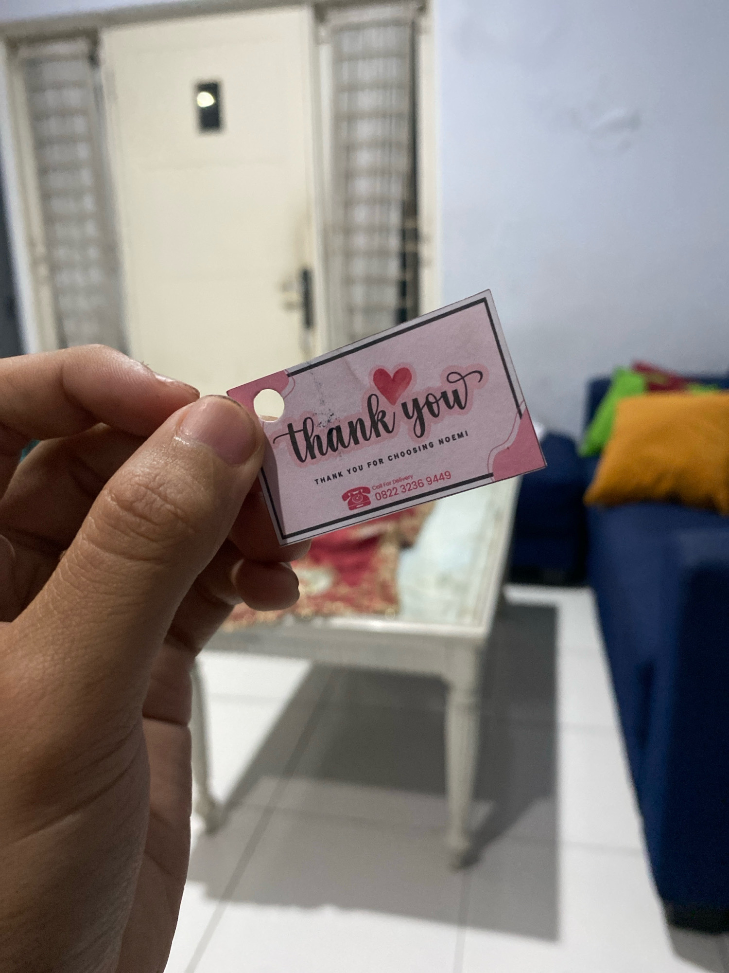 A person's hand is holding a pink thank you tag with "thank you" written in cursive, a heart, and a phone icon. Below, it says "THANK YOU FOR CHOOSING NOEMI" and "Call For Delivery 0822 3236 9449". The background shows a white door with a dark rectangular window in the center, a white table with ornate legs and a patterned cloth on top, and a dark blue sofa with orange and green cushions. The floor is tiled. This image was taken inside a residential building in Klurak, Indonesia.