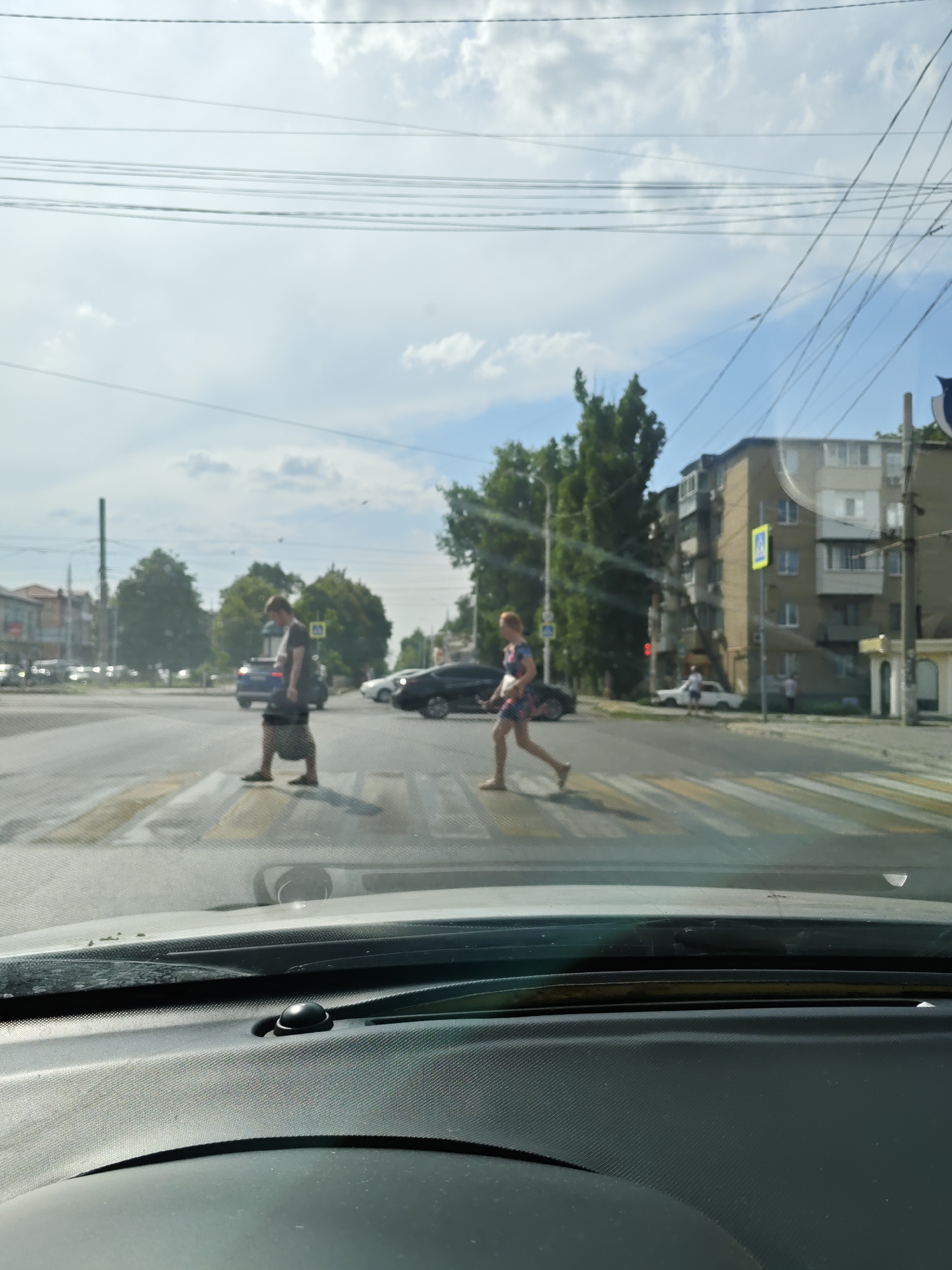 An outdoor, daytime, street-level view of a pedestrian crossing at an intersection in Novocherkassk, Russia. The image is taken from the perspective of someone inside a vehicle, looking out through the windshield, which is somewhat dirty and shows reflections. Two pedestrians are visible on the zebra crossing. On the left, a person wearing a mask, a dark t-shirt, and shorts is walking across the street. On the right, a person in a colorful dress is also crossing. Several cars are on the road, and a multi-story apartment building stands on the right side of the intersection. Telephone wires are strung across the sky, partially obscuring the clouds. The overall scene depicts a typical urban environment with everyday activities.