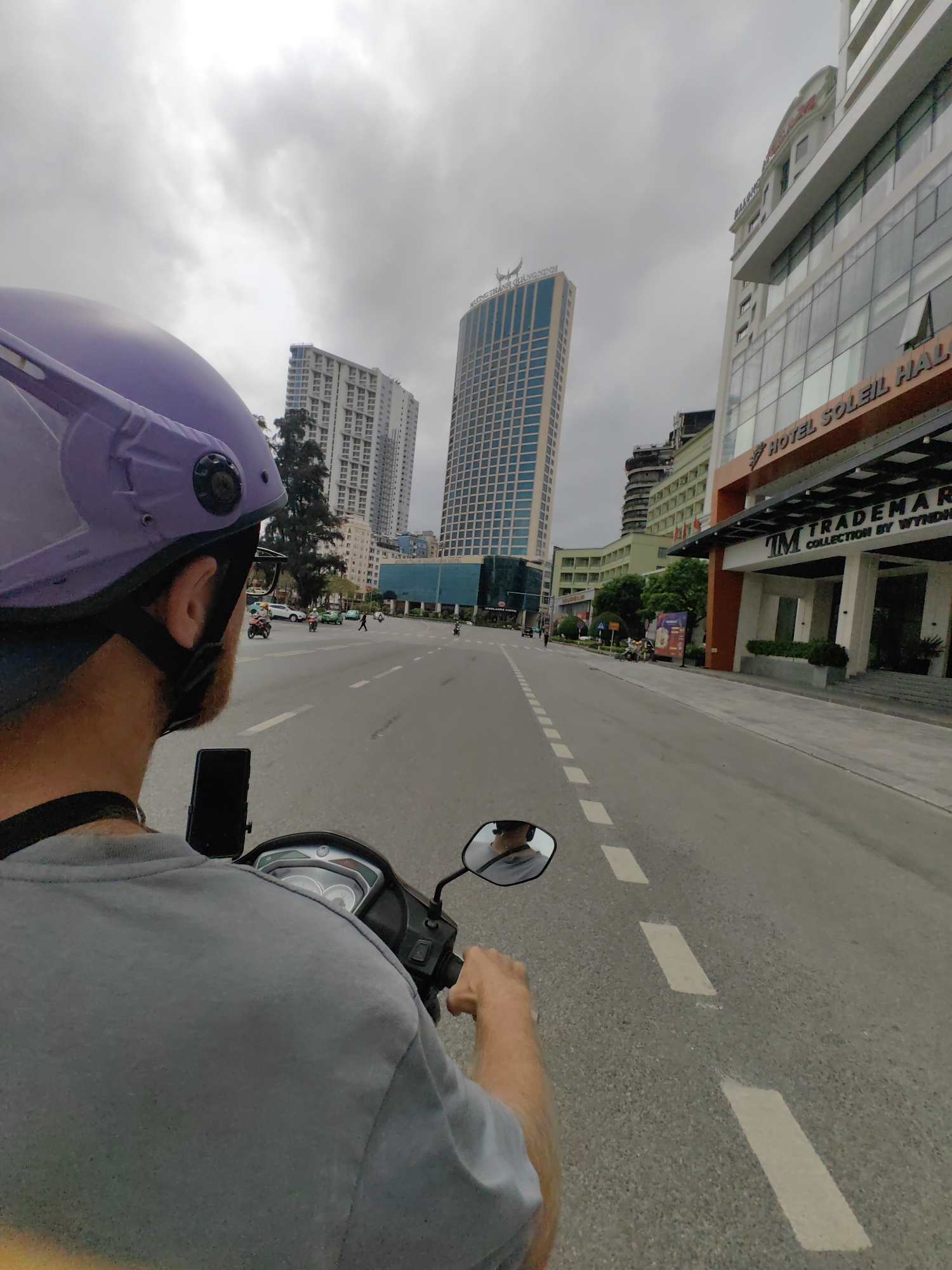 The image captures a first-person perspective from behind a person riding a scooter on a multi-lane urban road in Bai Chay Ward, Vietnam. The rider wears a purple helmet and a grey t-shirt. A smartphone is mounted on the scooter's handlebars, positioned behind the rider's left shoulder. The scooter's dashboard and a rearview mirror, reflecting a blurred view of the rider, are visible.
The sky is overcast and cloudy, suggesting a dull daytime atmosphere. The paved road features white dashed lane markings, guiding moderate traffic that includes other motorbikes and a white car in the distance.
Tall, modern buildings line both sides of the street. On the right, a prominent hotel building features signs reading "HOTEL SOLEIL HALONG" and "TRADEMARK COLLECTION BY WYNDHAM" above its entrance. To the left, a tall skyscraper displays "VƯƠNG THÀNH QUẢNG NINH" near its summit. Further down the street on the left, a "Highlands Coffee" sign is also visible. The scene depicts a bustling urban street environment with modern infrastructure, characteristic of a city in Vietnam.