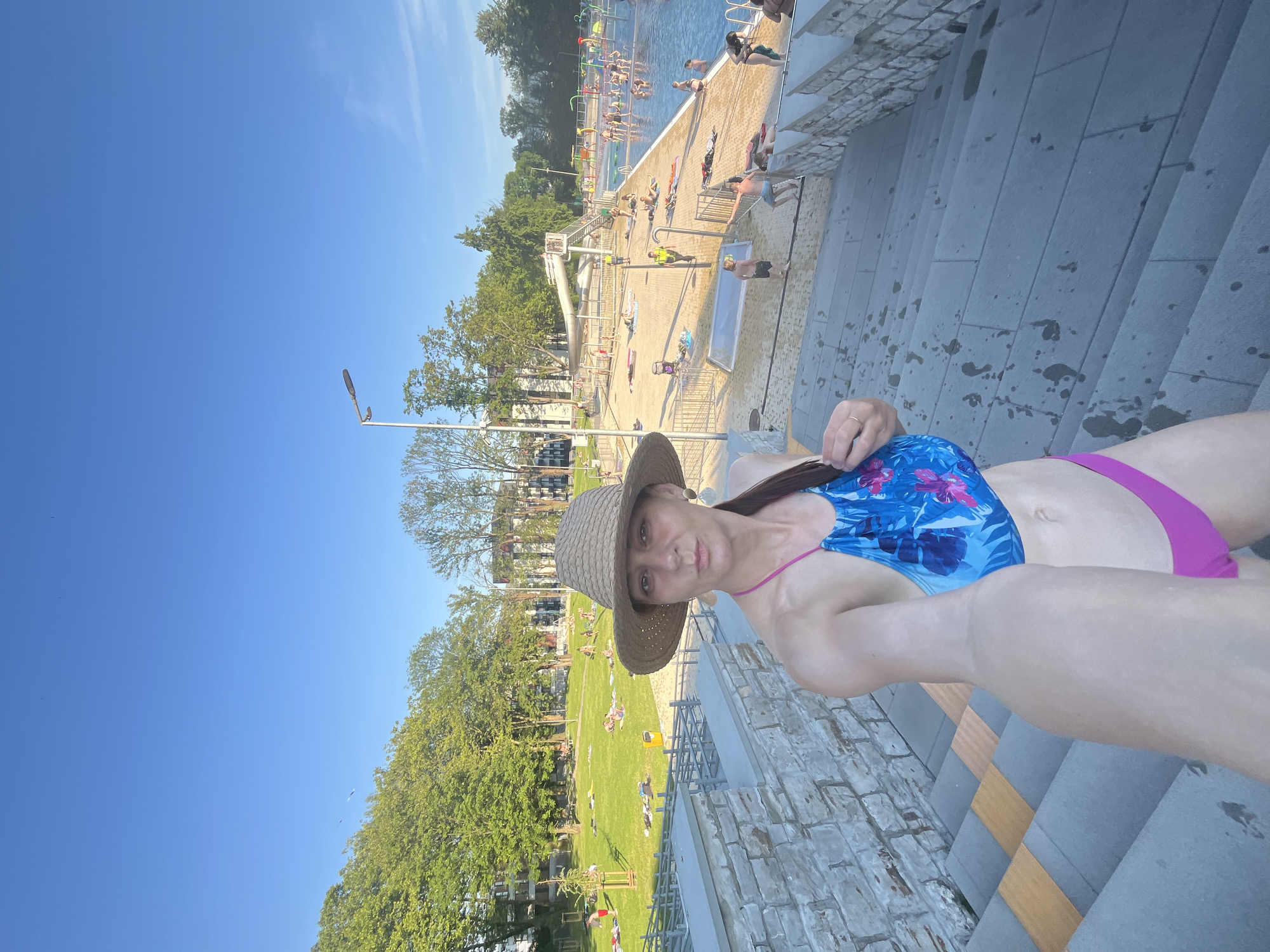 A woman in a straw hat and a blue and pink bikini poses for a selfie on a stone staircase at an outdoor swimming pool complex. She is looking at the camera with a neutral expression. Behind her, a large swimming pool is filled with people, some swimming and others lounging on the deck. A waterslide is visible in the background. The area is surrounded by trees and buildings, suggesting an urban park setting. The sky is clear and blue.