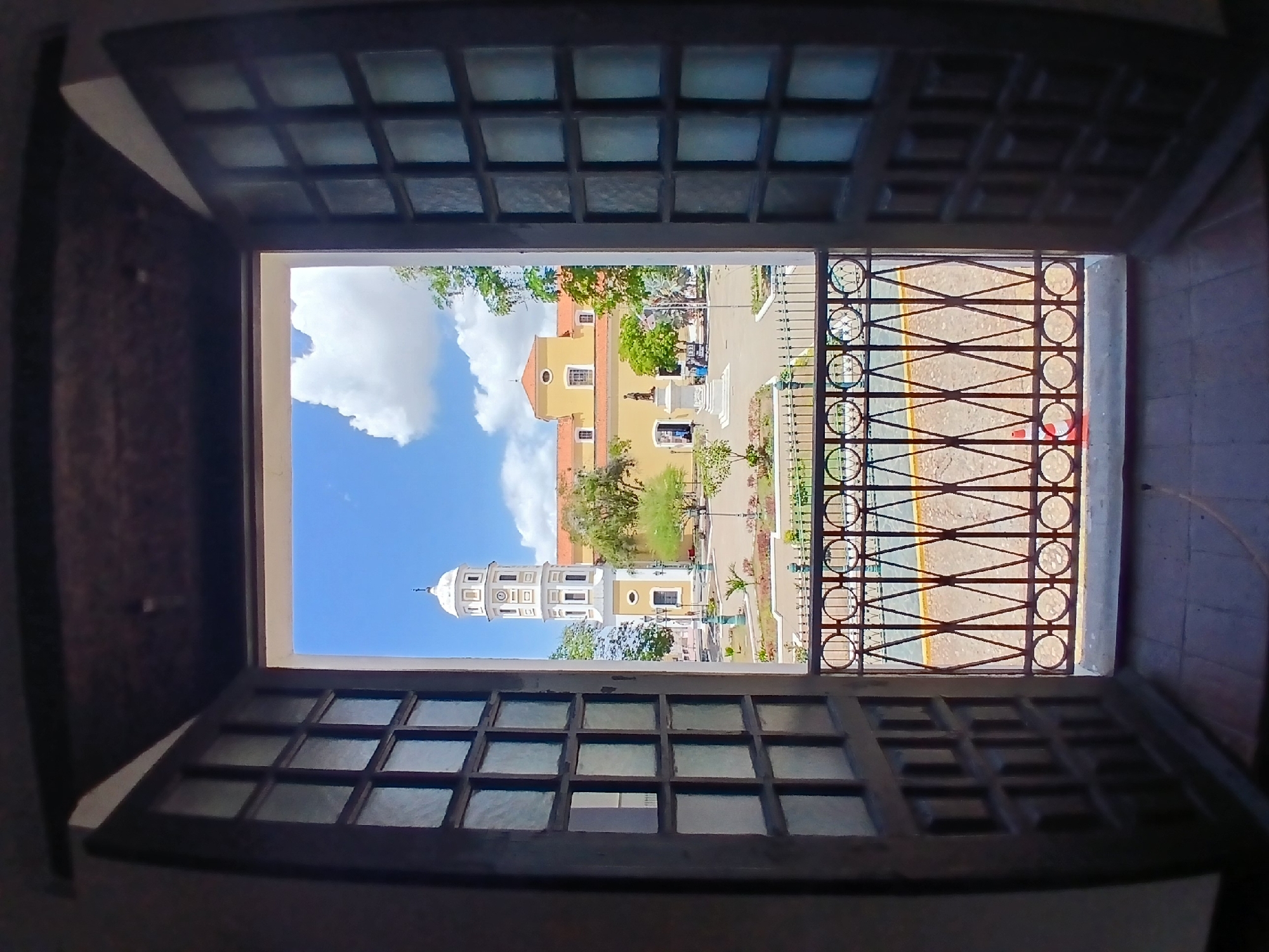 The image depicts a view through a window in Ciudad Bolivar, Venezuela, showcasing the Plaza Bolivar with its distinctive yellow buildings and white-domed tower. The window is composed of multiple small glass panes, framed by a dark-colored structure. A wrought-iron fence is visible outside the window. The Plaza features trees, paths, and a monument. A blue sky with white clouds serves as the backdrop.