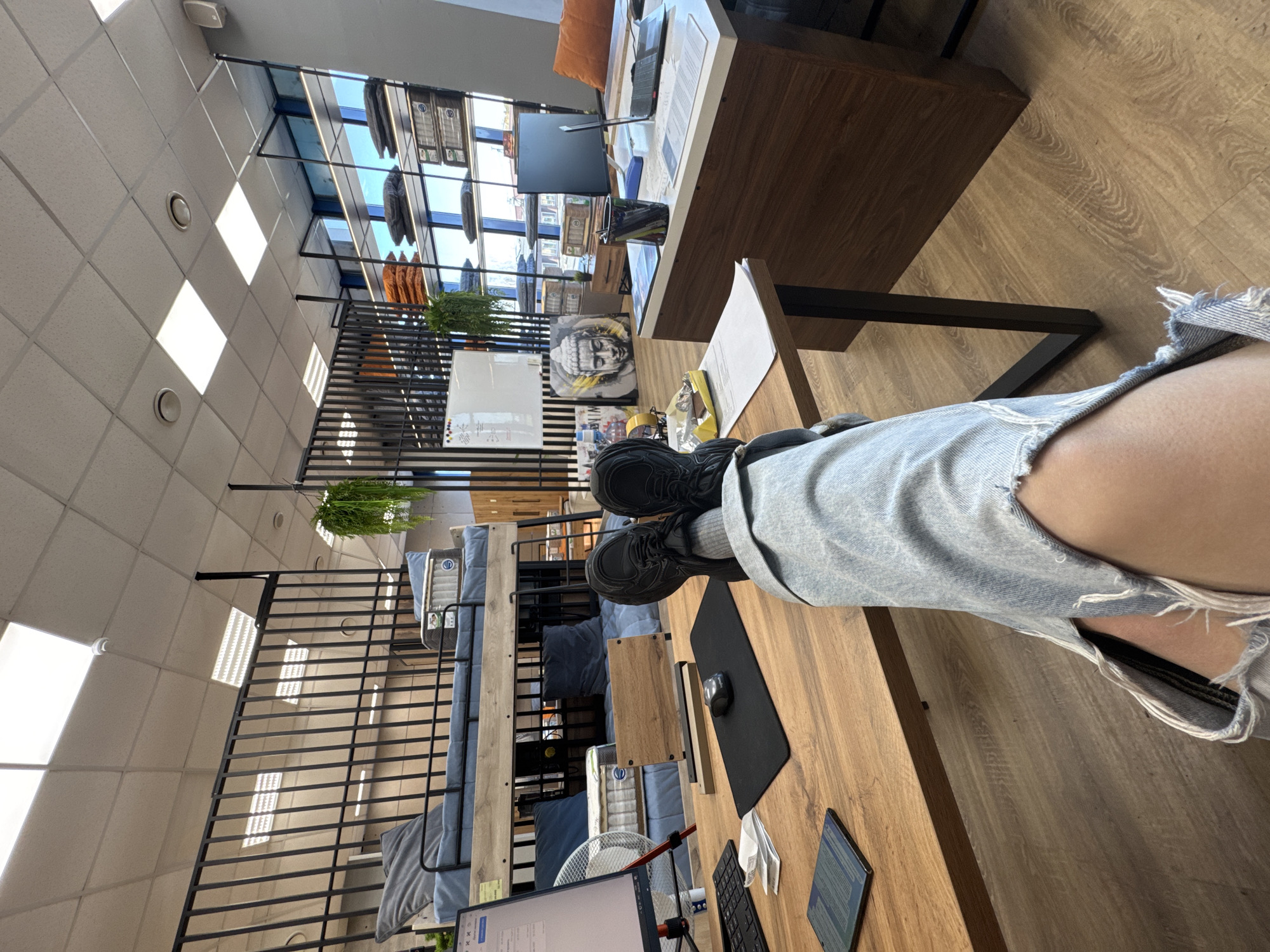 The image presents a first-person perspective from a seated position in what appears to be a furniture store. The individual's legs, clad in ripped light blue jeans and black sneakers with grey socks, are positioned atop a wooden desk. On the desk, there is a black mousepad with a computer mouse, a keyboard, a tablet, and a white fan.
The store features multiple levels accessible by stairs, where displays of beds with blue mattresses and pillows are presented. Overhead, there are hanging plants. A larger wooden desk with a computer monitor is visible in the background.
The location appears to be in Chaikovsky, Russia, evidenced by the storefront displays visible through the large windows showcasing various furniture items and designs, possibly indicating the merchandise offered at this store. A Buddha statue is also apparent.