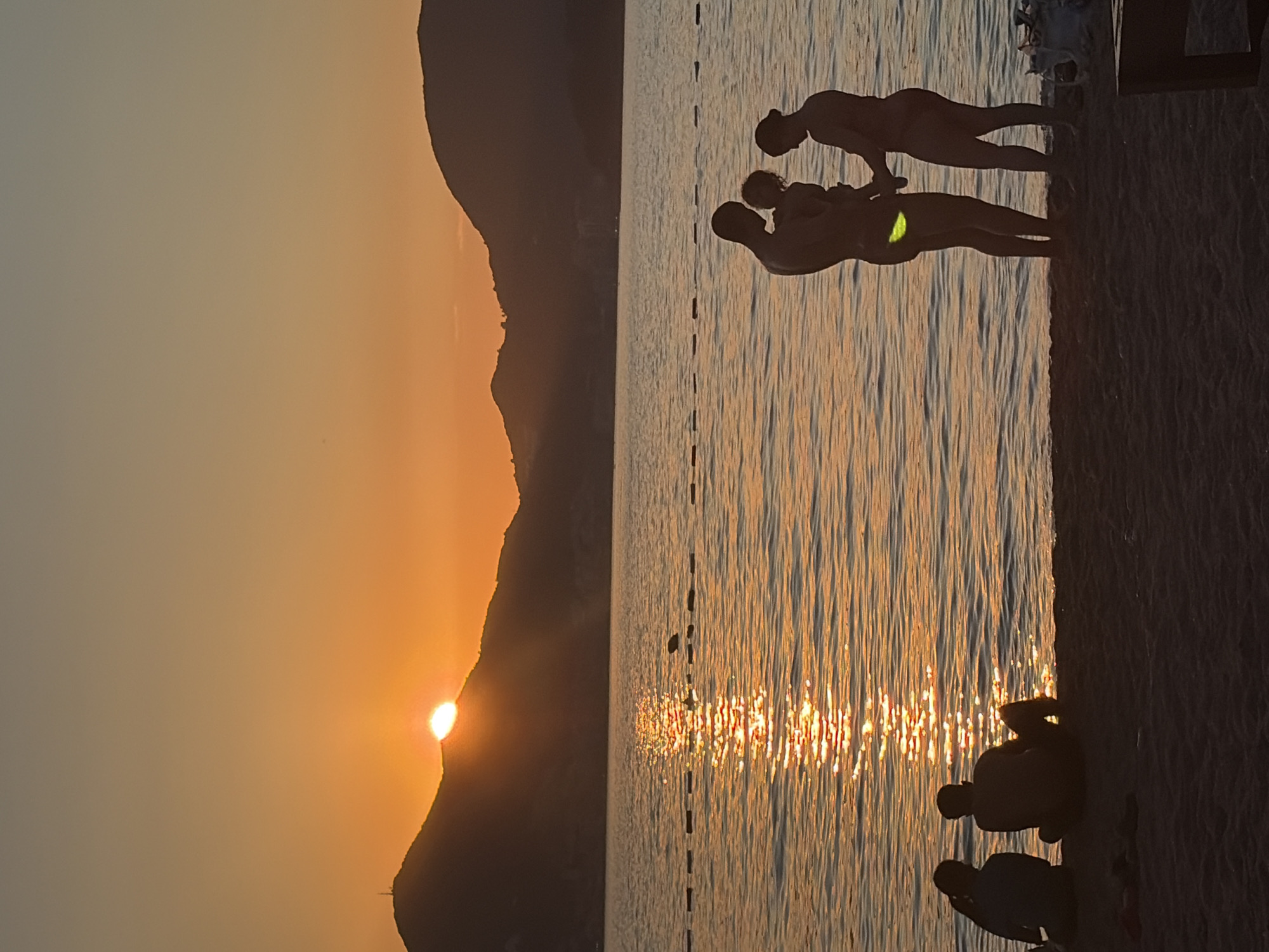 A group of people are silhouetted against a vibrant orange sunset over the sea at Sveti Stefan, Montenegro. The sun is partially obscured by a mountain range in the distance, casting a shimmering golden reflection on the water. On the beach in the foreground, two individuals sit facing away from the camera, while a family of three stands near the water's edge. The father holds a young child, and the mother stands beside them, her hand held by the father. The scene depicts a tranquil evening by the coast.