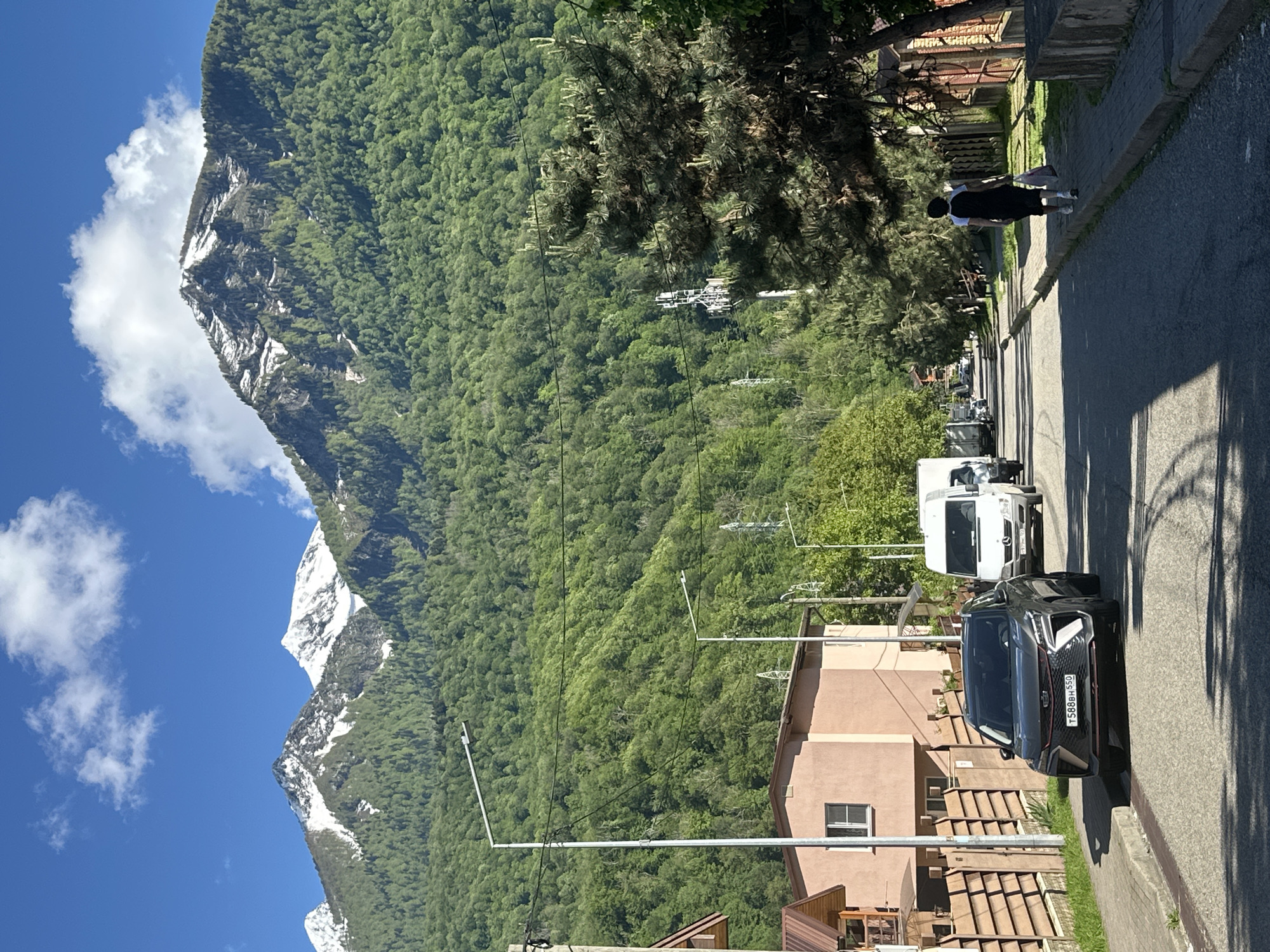 This media file captures a vibrant, sunlit scene in Krasnaya Polyana, Russia, set against a backdrop of majestic mountains. The left side of the image (when viewed upright) is dominated by towering mountains, densely covered in rich green forest. Several of the higher peaks are rugged and partially snow-capped, standing out under a clear blue sky adorned with scattered white clouds.
In the foreground and to the right, a paved street runs along what appears to be a residential area. A person, dressed in dark clothing and carrying light-colored bags, walks along the sidewalk, moving away from the viewer. Large trees, possibly pines, line the street, casting long shadows that indicate bright sunshine.
Several vehicles are parked on the street. Most prominently, a black SUV, which appears to be a Geely model, is parked close to the camera, displaying a Russian license plate "T588BH 550". Behind it, a white cargo van, resembling a Mercedes-Benz Sprinter, is also visible. Other vehicles are partially obscured further down the road.
Buildings with light brown or beige facades and tiled roofs line the street, suggesting residential or guesthouse structures. Electrical poles, streetlights, and overhead wires are present throughout the street level. In the mid-ground, nestled within the dense forest on the mountain slopes, sections of a ski lift or cable car system can be identified. The scene conveys a sense of a picturesque mountain town thriving amidst stunning natural beauty on a clear day.
