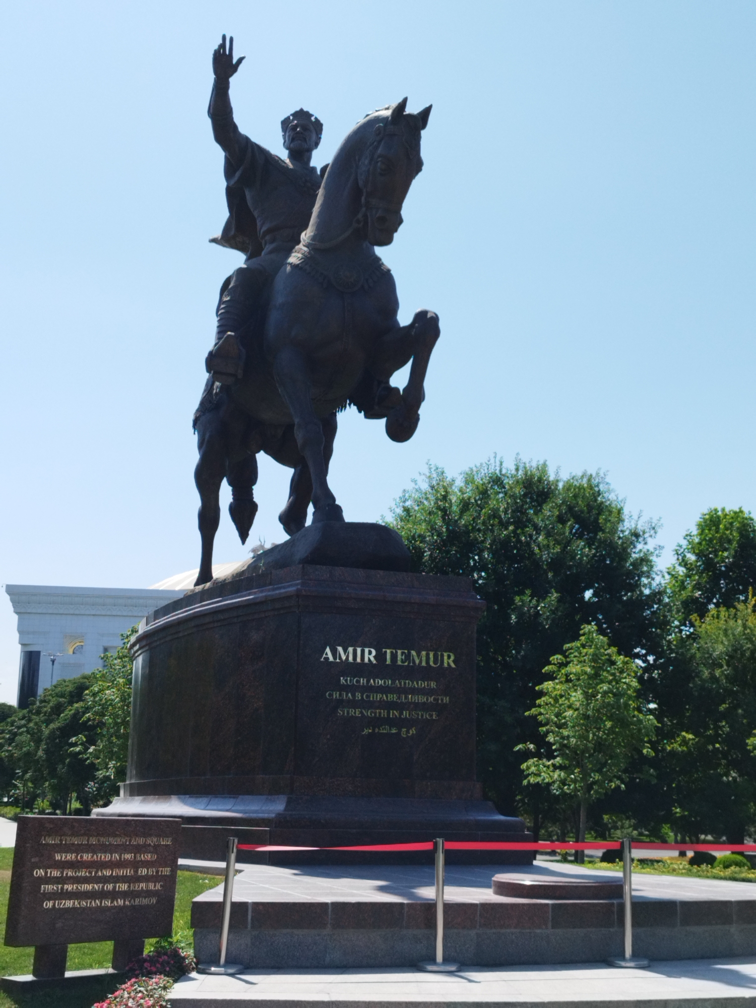 A bronze equestrian statue of Amir Temur, a historical figure, stands prominently against a clear blue sky. The statue depicts Amir Temur on horseback, with his right arm raised, a gesture of power or address. The horse is depicted in motion, with its front legs raised. The statue is mounted on a large, multi-tiered granite pedestal. Inscribed on the pedestal is "AMIR TEMUR" in large letters, followed by text in what appears to be multiple languages, including Russian and possibly Uzbek.
To the left of the pedestal, a smaller, separate granite monument is visible. This monument bears an inscription detailing the creation of the Amir Timur Monument and Square in 1993, based on a project initiated by the first president of the Republic of Uzbekistan, Islam Karimov.
Behind the statue and pedestal, lush green trees form a dense backdrop, suggesting a park or public square setting. In the far left background, a modern-looking white building with arched windows and a domed roof is partially visible. Red velvet ropes on silver stanchions cordon off the immediate area around the statue's base, indicating public access is restricted. The overall scene is well-maintained, with paved walkways and manicured greenery.