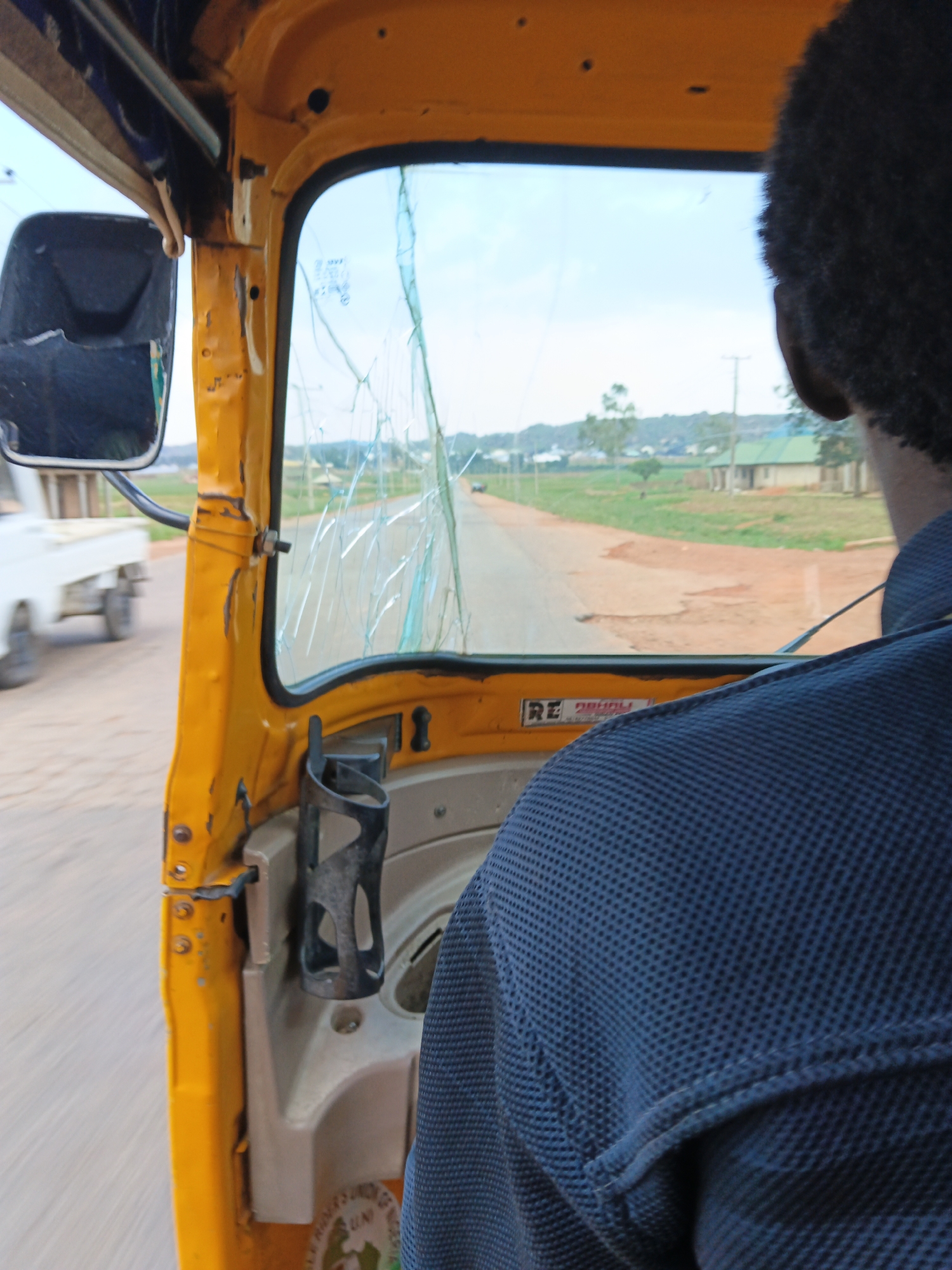The image shows the interior of a yellow, three-wheeled vehicle, commonly known as an auto-rickshaw or tuk-tuk, viewed from the perspective of a passenger. The driver's head and shoulder, wearing a dark blue patterned shirt, are visible on the right side of the frame, looking forward through the windshield. The windshield is cracked, with multiple spiderweb fractures across its surface. A black side-view mirror is attached to the left of the vehicle's interior. The dashboard area is visible, featuring a dark colored cup holder. Outside the vehicle, a paved road is seen, with a blurry white truck in motion on the left side. The landscape beyond the road appears to be rural or semi-rural, with fields, scattered buildings, and trees under a cloudy sky. The scene suggests the vehicle is in motion.
