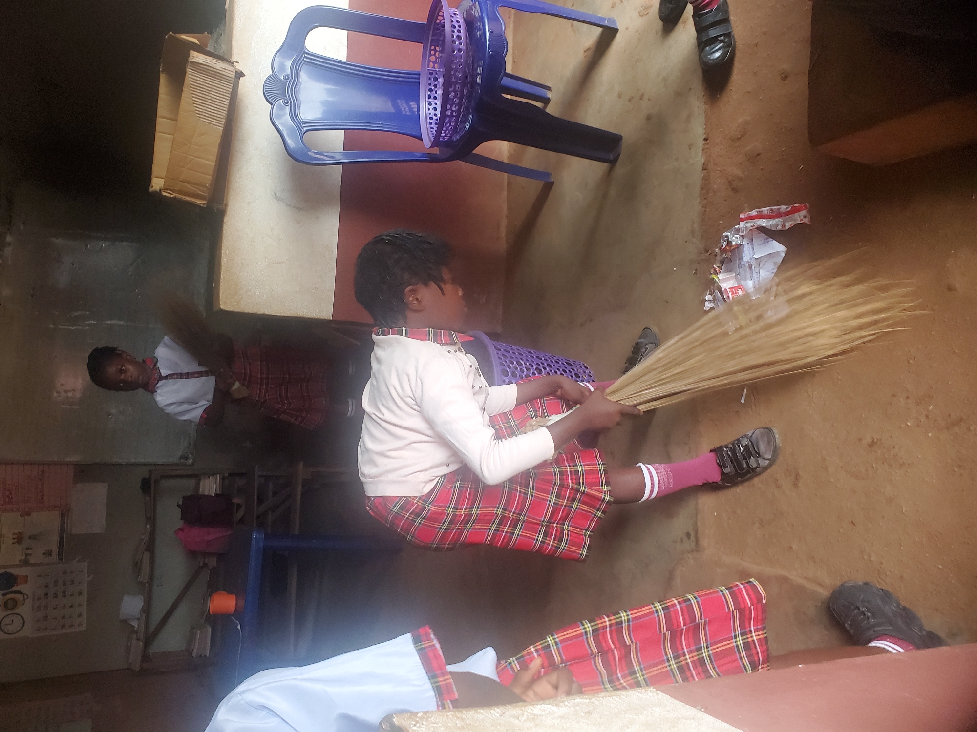 A young person in Ukwu-Abwa, Nigeria, sits indoors on a blue plastic basket, holding a bundle of sticks. The person wears a white long-sleeved top, a red plaid skirt, and pink socks. To the left is a second young person in a similar uniform standing near a calendar. A blue plastic chair and a cardboard box sit nearby. Debris is on the floor.