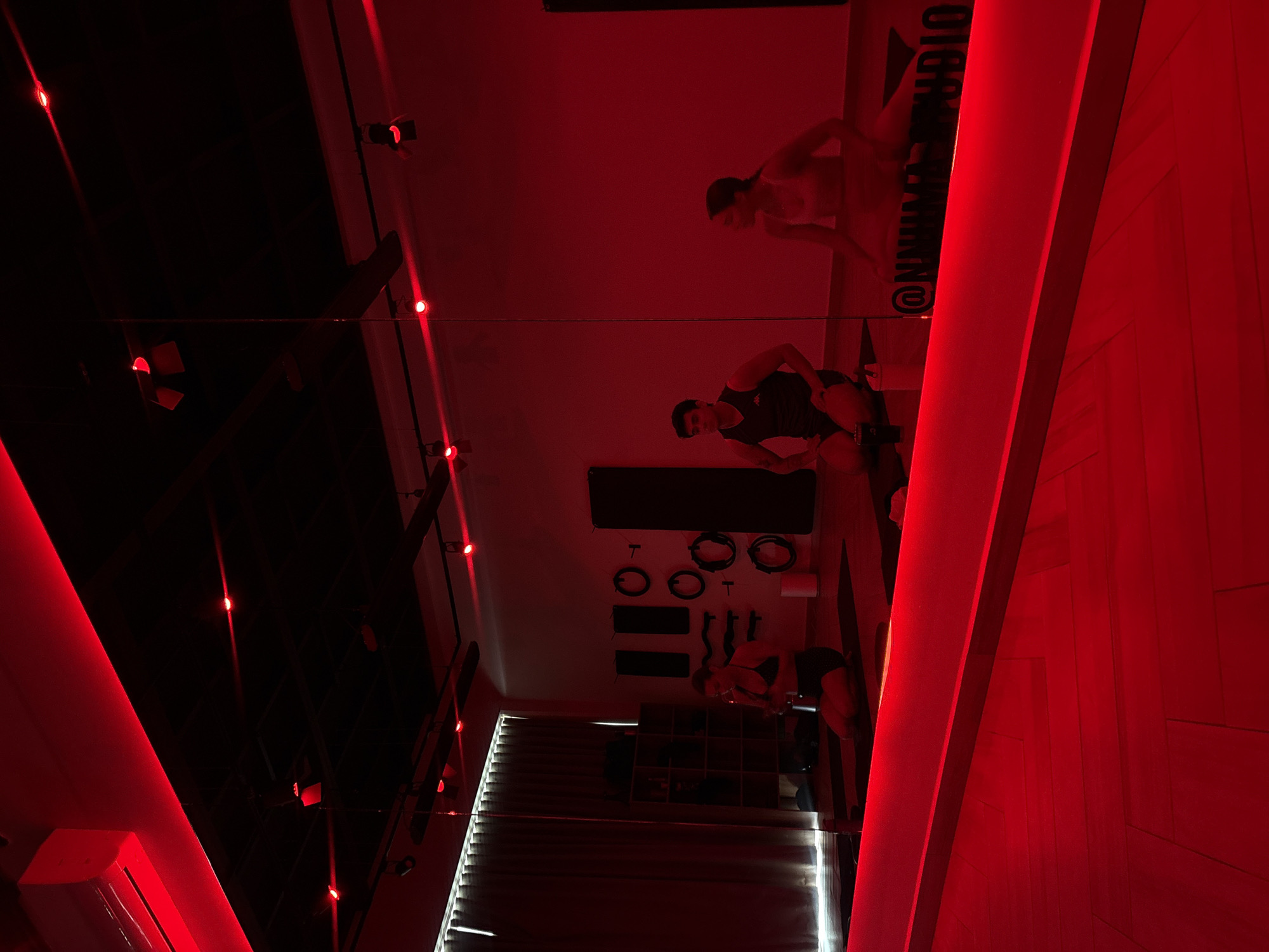 The image depicts the interior of what appears to be a gym or workout studio in San Bernardino Tlaxcalancingo, Mexico, characterized by its predominant red lighting. There are at least three individuals present, engaged in fitness activities. One person is visible near the top of the frame, and another is situated closer to the center. A third individual can be seen further back in the scene near equipment. The studio's equipment includes various weights, and a mirror. The architecture includes a slatted window covering, presumably to control light. The lighting system consists of recessed lights and LED strip lights.