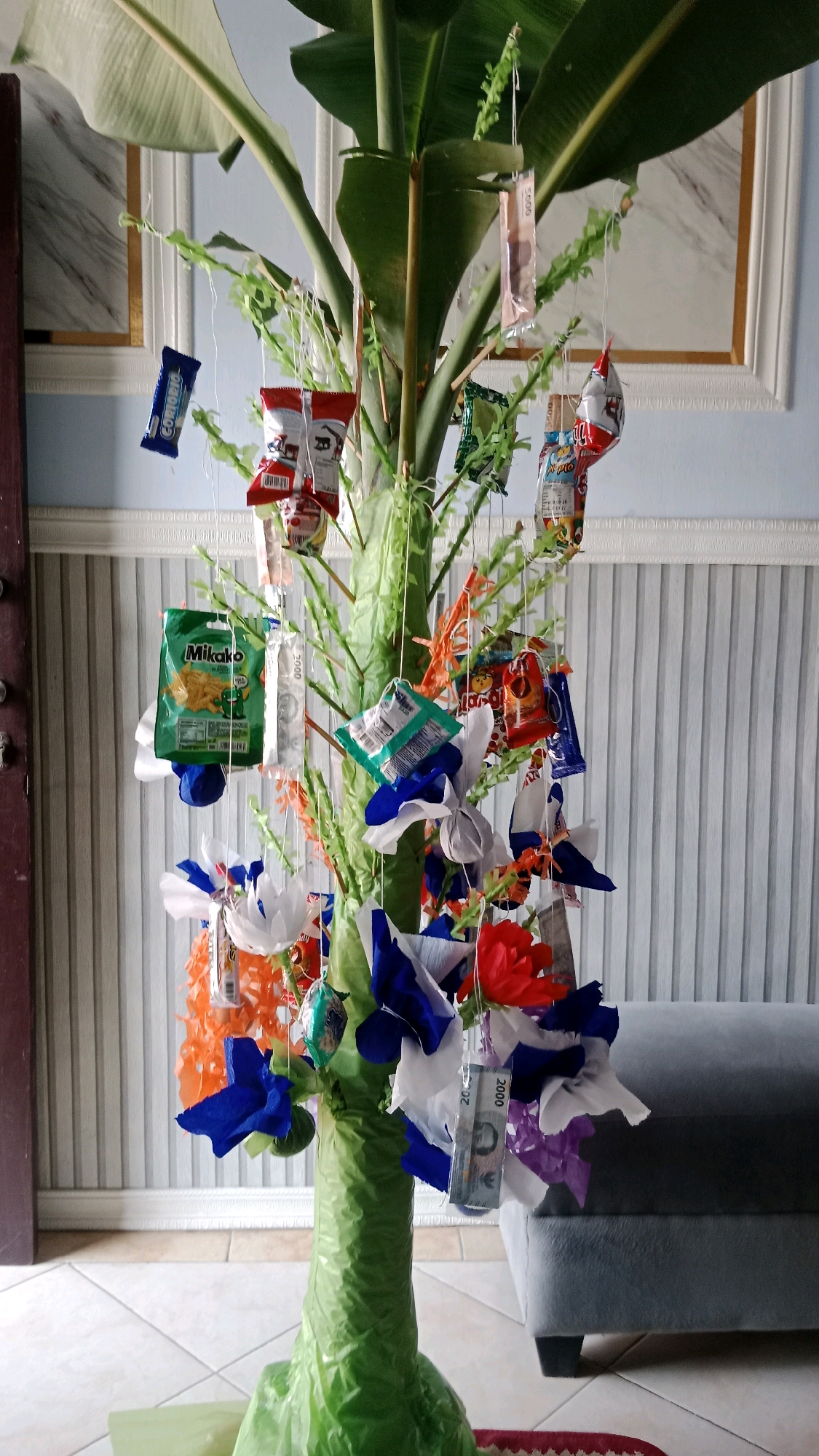 A tall, green tree-like structure is decorated with small packets of snacks, colorful paper flowers, and Indonesian currency. The tree is wrapped in green plastic, and its base is also covered in green plastic. The backdrop is a wall with vertical white paneling and a framed picture with a gold border. A gray sofa is visible to the right of the tree. The floor is tiled in a checkered pattern. The overall scene appears to be an indoor celebration or decoration.