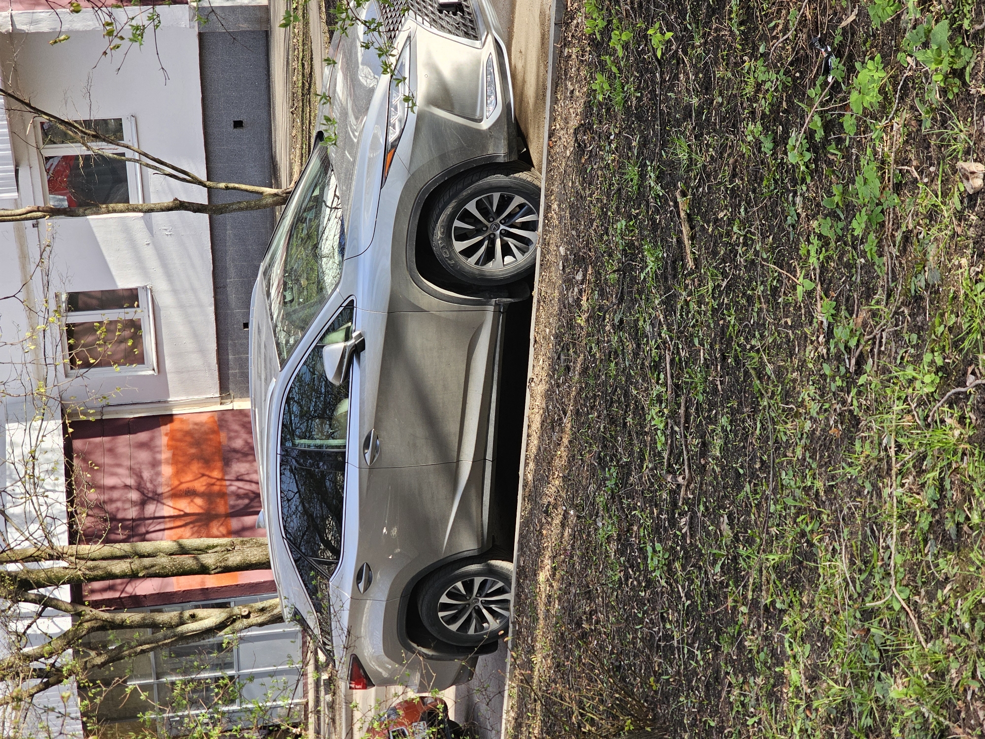 The image, when viewed in its correct orientation, depicts an outdoor scene in Khimki, Russia, featuring a parked car adjacent to a building and a patch of natural ground.
The primary subject is a light-colored SUV, appearing to be a Lexus RX model, distinguishable by its front grille and the Lexus logo visible on the wheel hubcaps. The vehicle is parked with its left side near a paved surface, and its right-side wheels are on an unpaved area of dirt and sparse, nascent grass.
In the background, to the left of the car, stands a light-colored, possibly white, multi-story building. It features several windows with white frames. A noticeable section of the building's lower wall or perhaps a garage door is painted in hues of orange and reddish-brown. A prominent tree with mostly bare branches or very early spring foliage stretches horizontally across the scene, partially obscuring the building and casting shadows.
The ground area to the right of the car is an unmanicured patch of dark soil interspersed with green sprouts, dry twigs, and sparse grass, suggesting the early stages of spring growth. A dark gray paved walkway or narrow road runs parallel between the car and the building.
The scene is illuminated by bright natural daylight, indicating a clear day. There are no people visible, and no specific activity or event is taking place beyond the car being parked. The overall atmosphere is calm and ordinary, characteristic of a quiet urban or suburban setting. The only visible text is the Lexus logo on the car's wheels.