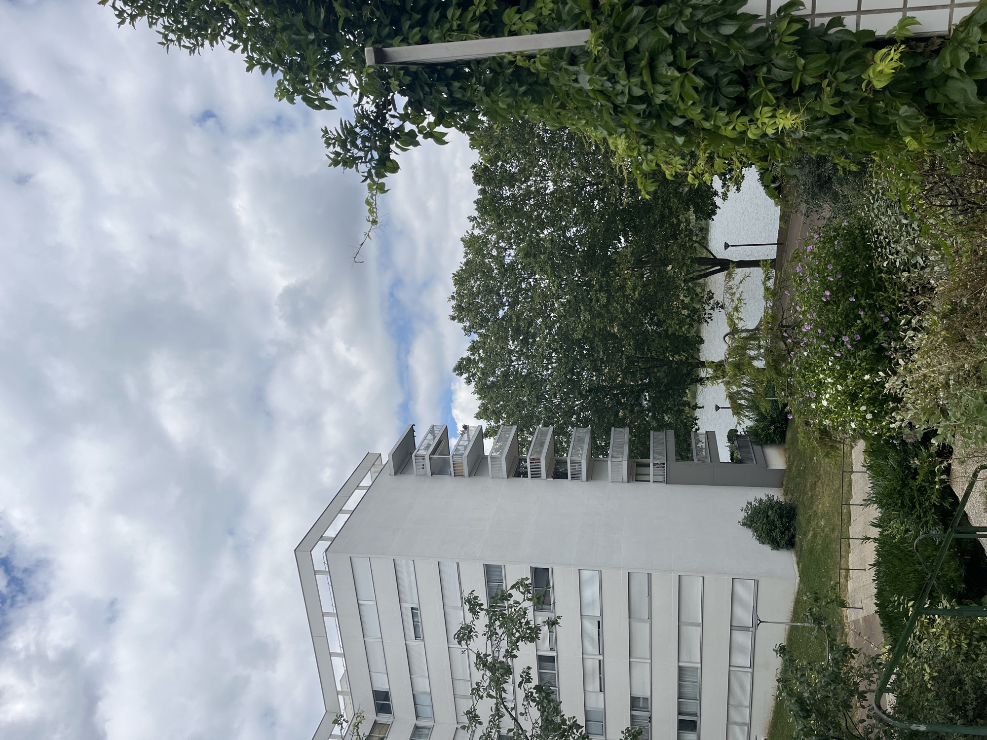 The image depicts a view of a residential building and its surroundings in Créteil, France. The building is a modern, white, multi-story apartment complex with balconies on one side. Large trees with dense green foliage are visible in the middle ground, with a body of water, likely a lake or river, behind them. The foreground features various plants and greenery, including flowering bushes and climbing ivy on a white tiled structure. The sky is overcast with a mix of white and gray clouds. The scene appears to be a residential area with green spaces and water features, characteristic of certain neighborhoods in Créteil, such as those near the Lac de Créteil. There are no people visible in the image.