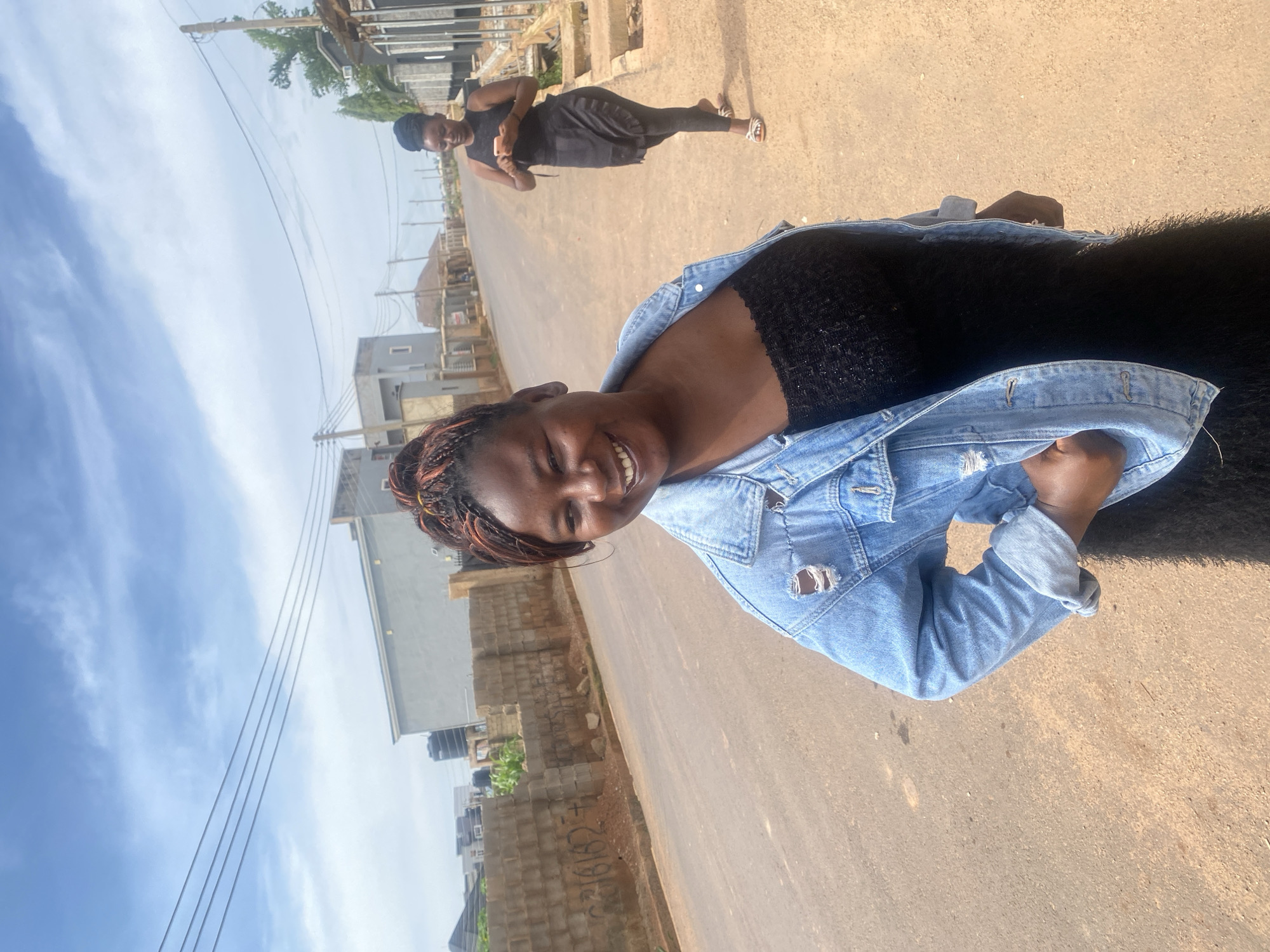 The image depicts an outdoor scene on a paved road in Bar Jirgi Summa, Nigeria. In the foreground, a young woman with braided hair is smiling and looking at the camera. She is wearing a distressed denim jacket over a black, textured top and dark-colored pants. Her left hand is placed on her hip. In the background, another young woman is walking away from the camera, looking down at her phone. She is wearing a black sleeveless top and dark leggings, with a black skirt or wrap over her leggings and a pair of sandals. The environment includes a dirt shoulder along the road, brick walls, some uncompleted buildings, and utility poles with overhead wires, suggesting a developing urban or suburban area. The sky is partly cloudy.