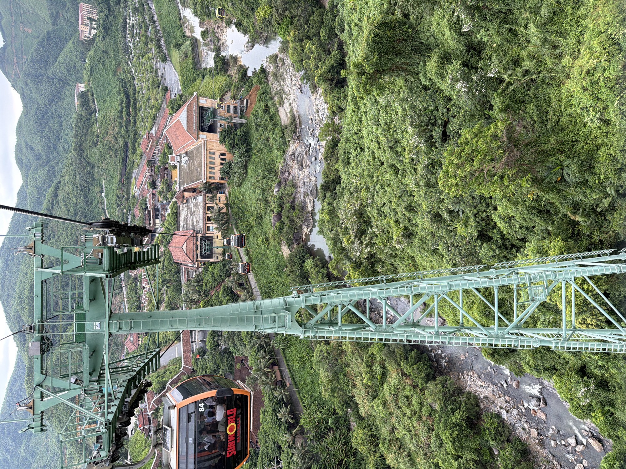 The image shows a view from a cable car in the Ba Na Hills area of Đà Nẵng, Vietnam. The green cable car structure is prominent in the foreground with an orange Sun World cable car visible. Buildings with red-tiled roofs are visible below among dense green vegetation. A river flows through the landscape.
