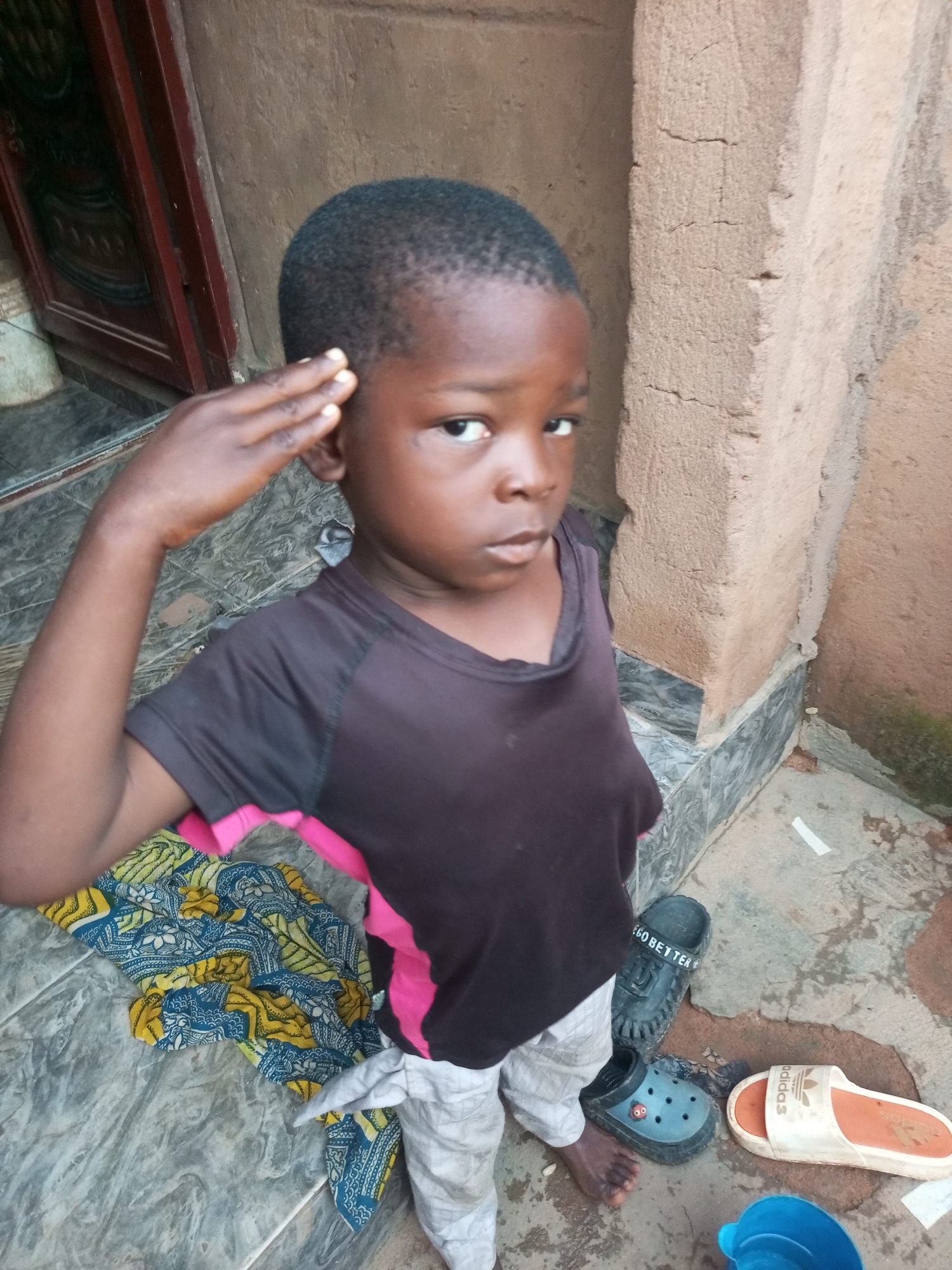 A young boy stands in an outdoor courtyard in Tempure, Nigeria. He is wearing a dark t-shirt with pink accents and light-colored pants. His right hand is raised to his forehead in a salute gesture. The ground is tiled and a colorful cloth is laid out on the left. Several pairs of shoes, including clogs and flip-flops, are visible on the ground near him, along with a blue bucket. The background consists of a rough, textured wall and a wooden door. The lighting suggests it is daytime.