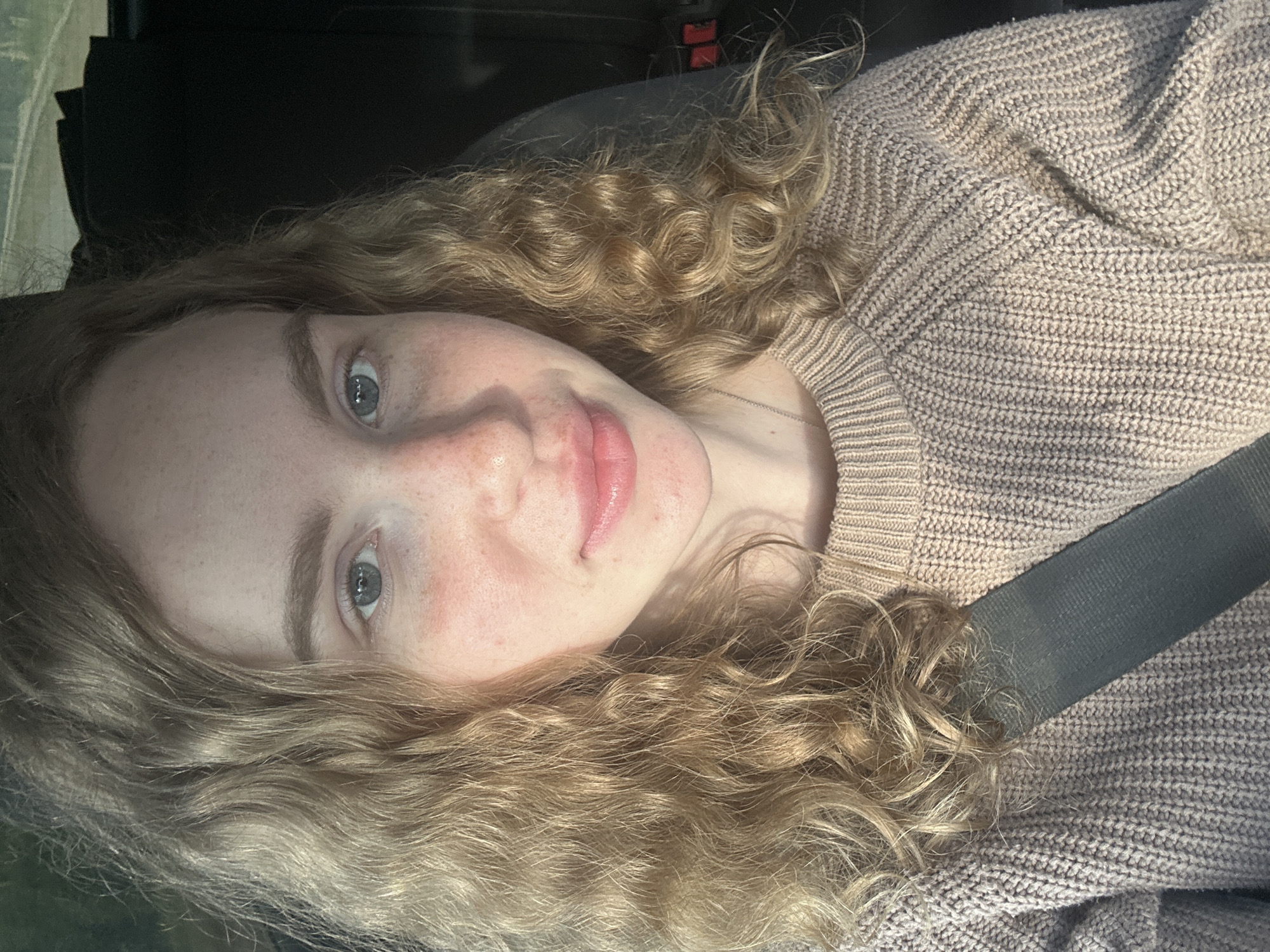 A selfie of a light-skinned person with curly blonde hair and light blue eyes wearing a beige knitted sweater and a gray seatbelt. The background appears dark, suggesting the interior of a vehicle. The location in the city of Vasilyevo, Russia, is undetermined from the image.