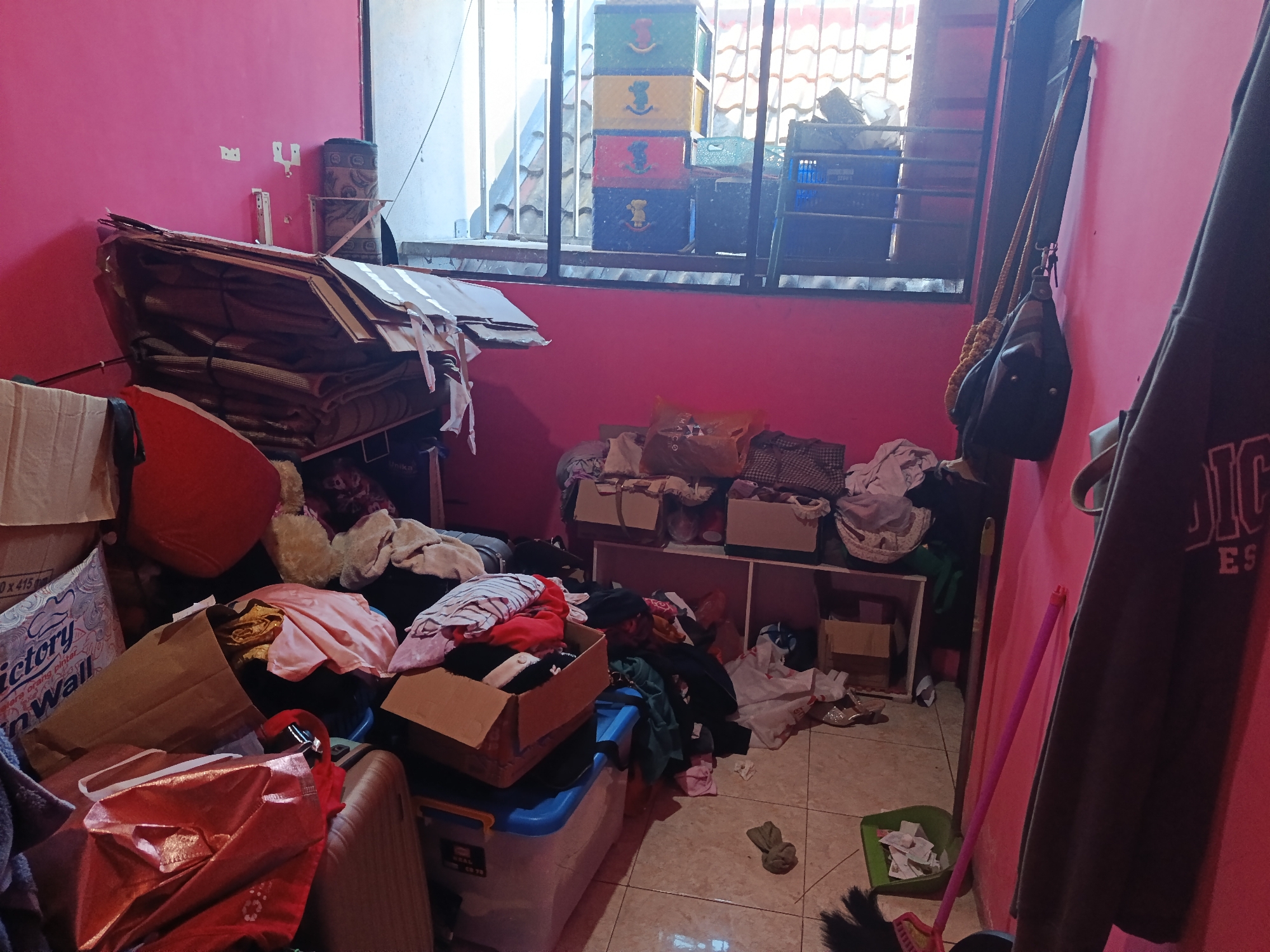 The image depicts a cluttered room with pink walls, likely a storage area or living space in a residential building in Keboananom, Indonesia. The room is filled with various items, including stacks of cardboard boxes, folded fabrics, and plastic containers. A large pile of what appears to be folded bedding or mattresses is leaning against the left wall. To the right, a window overlooks an outdoor area with tiled roofing and what seems to be a clothesline. There are also two bags hanging on the wall near the window, and a dark-colored garment is draped over the right side of the frame, obscuring part of the wall. The floor is tiled and strewn with miscellaneous objects, including a green dustpan with some papers inside and a broom with a pink handle. The overall impression is one of disarray and significant accumulation of household items.