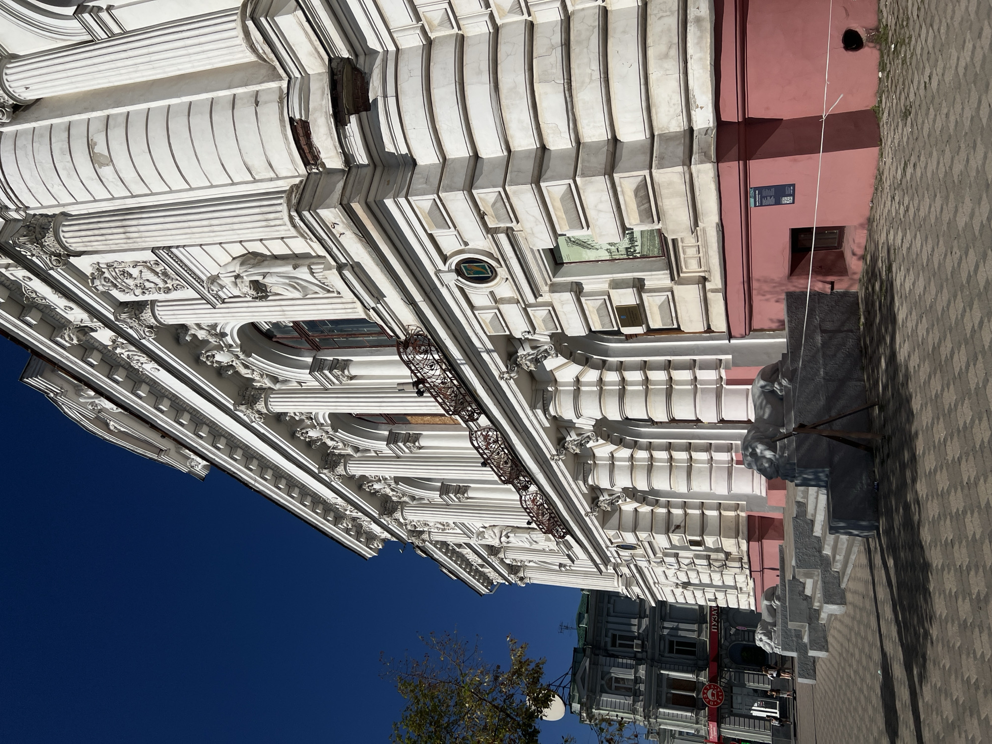A low-angle, wide shot captures the ornate facade of a white, classical-style building against a clear blue sky in Kharkiv, Ukraine. The building features elaborate architectural details including columns, sculptures, and decorative carvings. A series of steps with two lion statues flank the entrance. To the left, a darker, more utilitarian building is partially visible. Paved ground with shadows from trees and the building stretches across the foreground. A few individuals are walking on the sidewalk in front of the darker building. The overall scene suggests an urban environment with historical architecture.