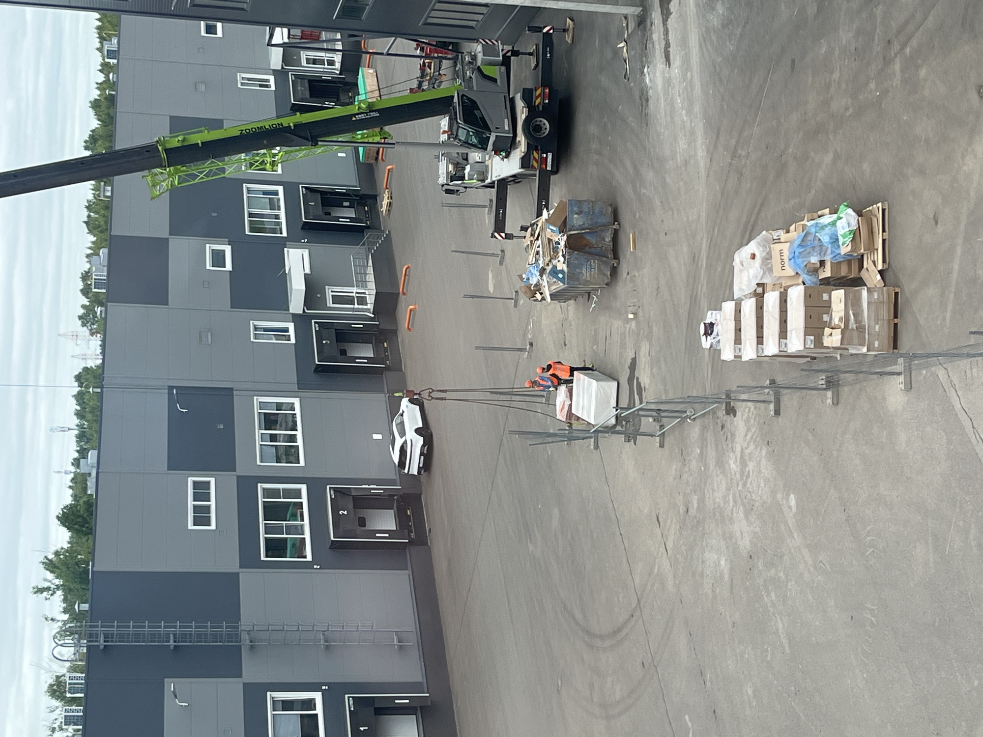This image captures an active industrial or construction site from an elevated perspective, likely on an overcast day. The main focus is a large, green and black "ZOOMILION" crane in operation, lifting a substantial metal frame or cage structure.
Two construction workers, identifiable by their orange safety vests and hard hats, are visible near the suspended load, appearing to guide or manage the lifted materials. The ground area is a large paved surface, showing tire marks and some debris. Near the crane's base, two blue dumpsters are filled with what looks like construction waste. Further right, multiple pallets are stacked with various boxes, some white and some brown cardboard. One box clearly displays the word "norm". A white car, resembling a Tesla Model 3, is parked closer to the buildings.
The surrounding structures are modern industrial buildings, featuring grey and black paneling, multiple windows, and several loading bay doors, some marked with numbers like "1" and "2". A metal ladder or walkway is affixed to one of the building facades. In the distant background, beyond the buildings and trees, power lines or towers are visible against a bright, cloudy sky, indicating a daytime scene. The overall image depicts a methodical material handling operation at an industrial facility in Zelenodol'sk, Russia.