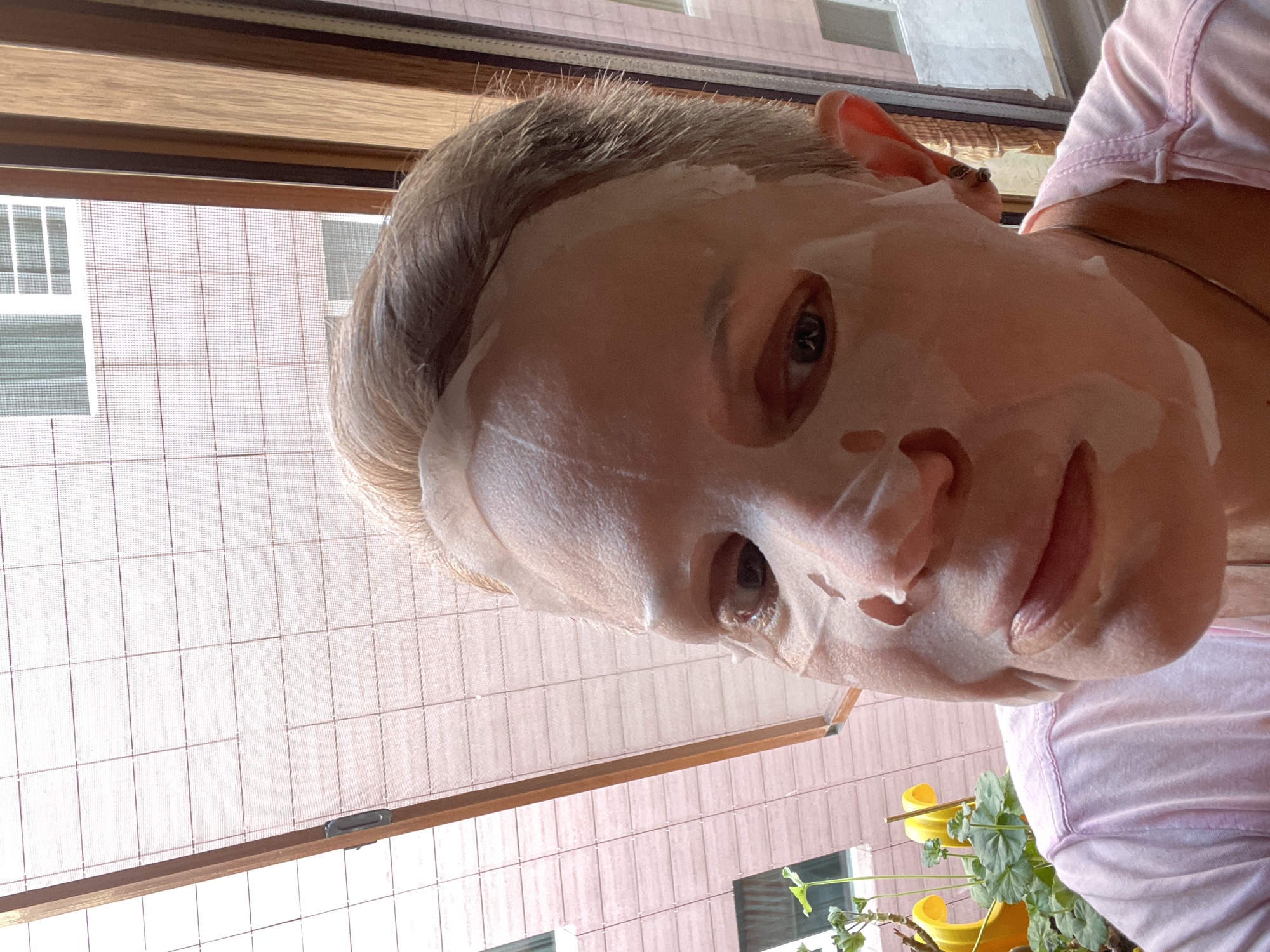 The image is a close-up selfie of a person with short blonde hair wearing a sheet mask. The person is wearing a pink t-shirt and a silver necklace. In the background, there is a window with wooden frames, a screen, and a building with pink brickwork. There are also some green plants visible on the left side of the image. The scene appears to be indoors near a window.