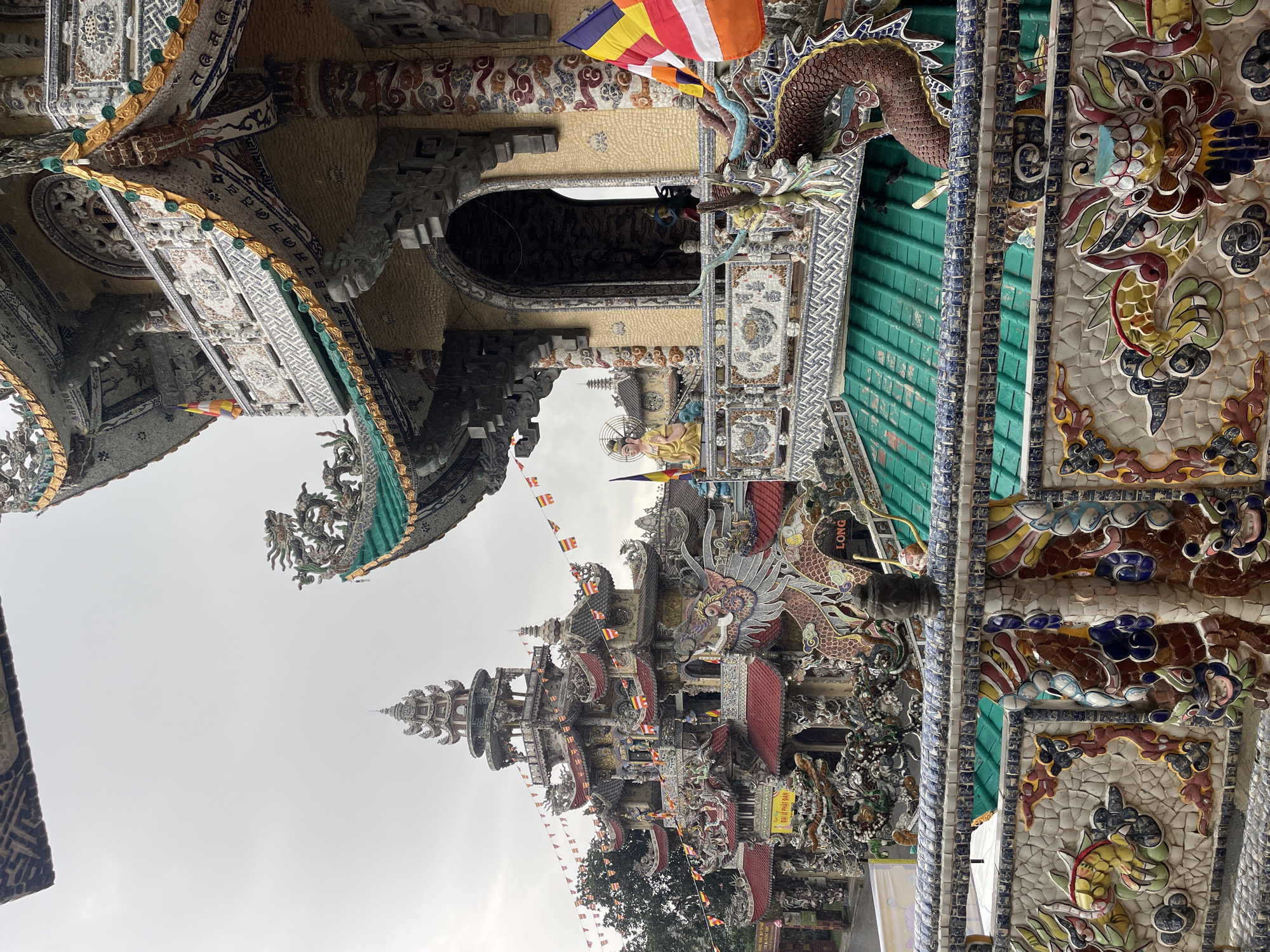 The image depicts the Linh Phuoc Pagoda, located in Xuân Thành, Vietnam. The structure is highly ornate, featuring intricate mosaics of glass and pottery shards. Architectural details include multiple roofs, spires, and dragon sculptures. Buddhist flags are visible, and the sky is overcast.