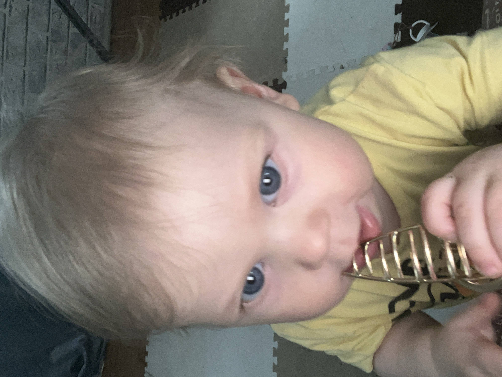 A close-up, rotated image shows a young child with blonde hair and blue eyes, wearing a yellow shirt. The child is holding and chewing on a gold-colored, cage-like object. The background includes a wall and a floor with interlocking foam tiles. There is no information related to Vesyoloye, Russia.