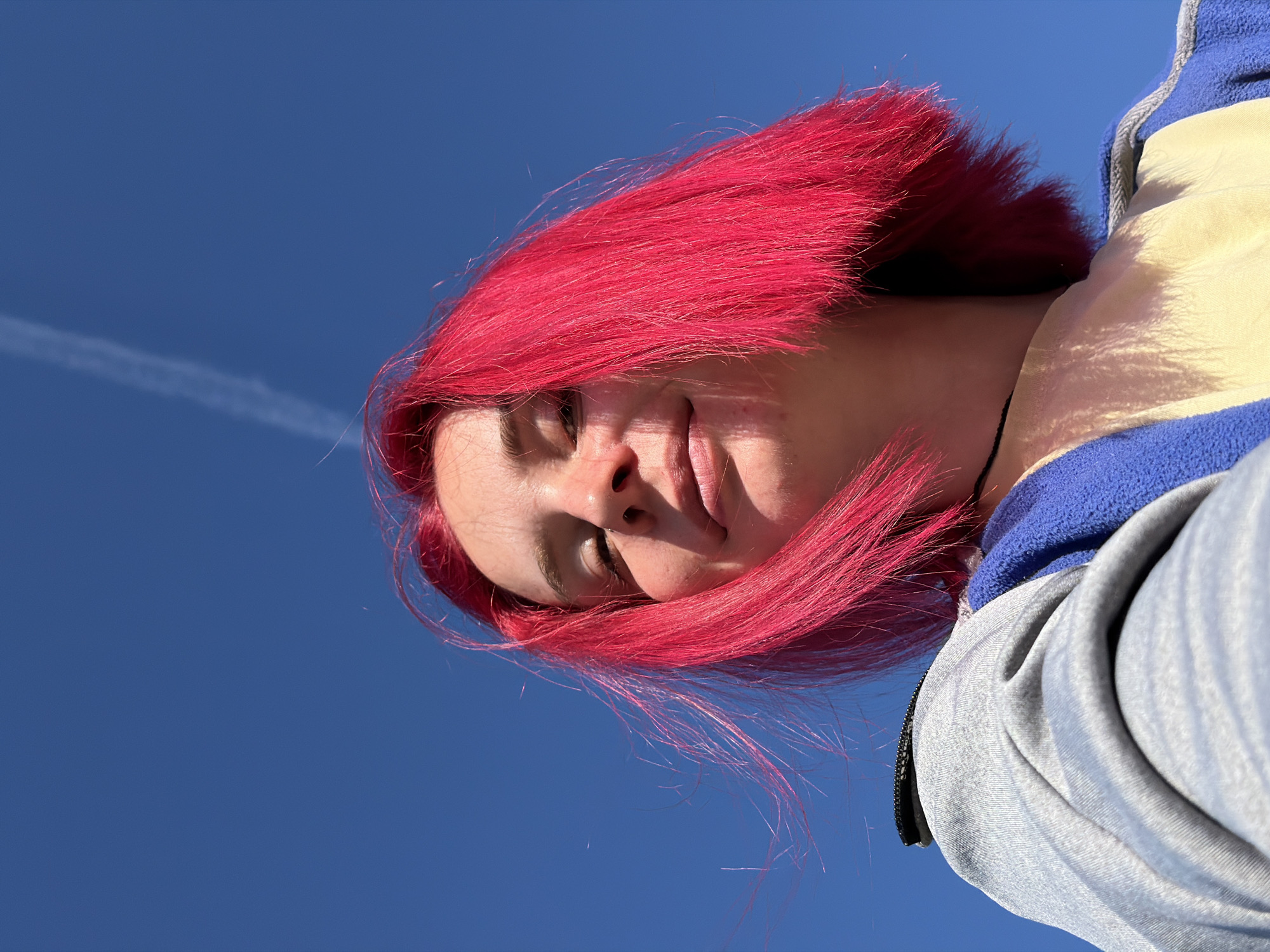 A low-angle, close-up shot captures a young person with bright pink hair against a clear blue sky. The individual is wearing a light yellow t-shirt and a blue and grey hooded jacket. Sunlight illuminates their face, casting shadows and highlighting a slight smile. A faint white contrail stretches across the sky in the upper right portion of the frame. The image appears to be taken outdoors, with no specific city landmarks visible to identify its location. The scene is static, with no discernible action taking place.