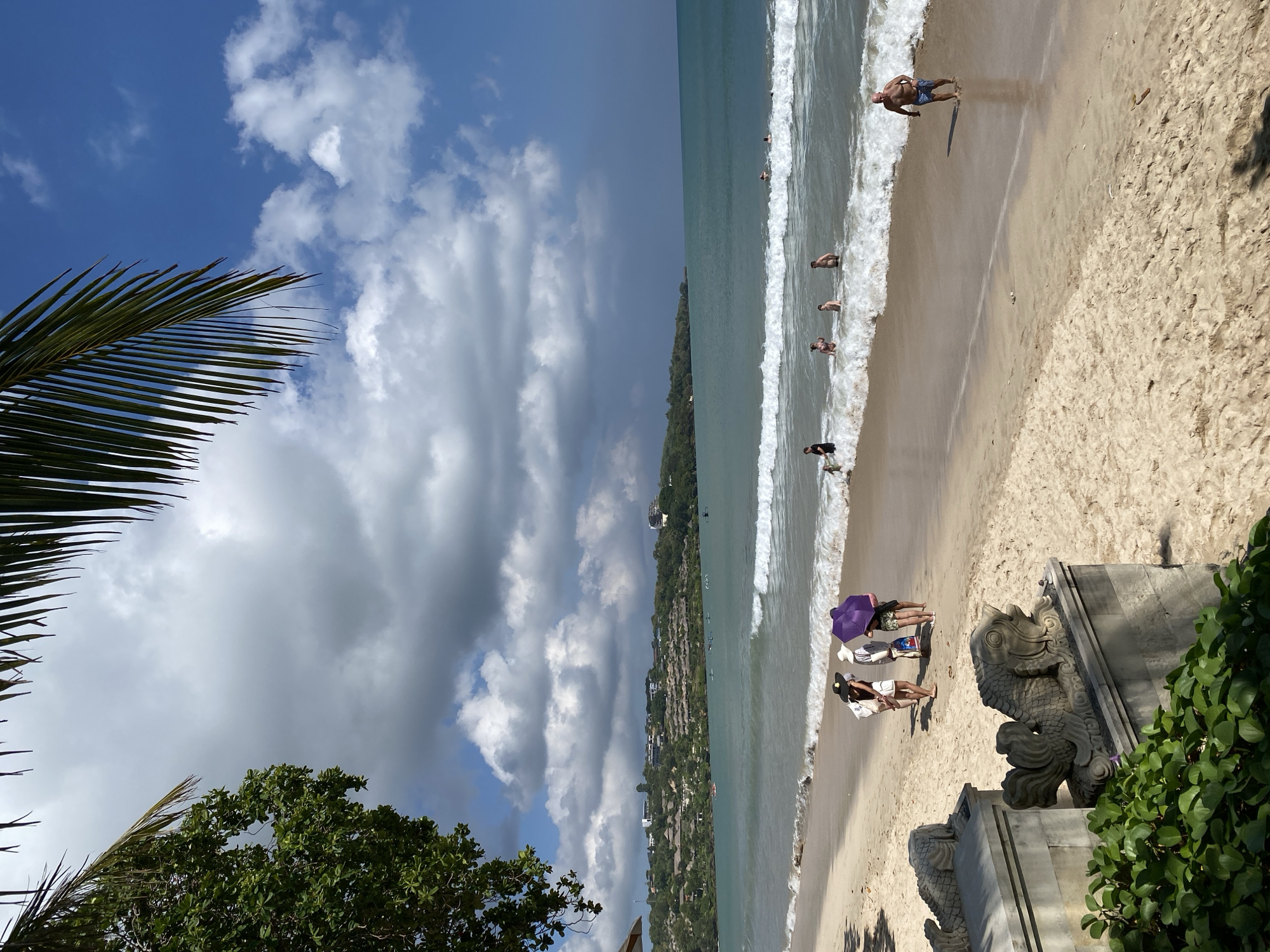 This image depicts a lively beach scene in Kuta Selatan, Indonesia, captured from a slightly elevated vantage point. The sky overhead features a mix of bright blue patches and white cumulus clouds, with some darker grey clouds contributing to a partially cloudy atmosphere. A large palm frond arches into the frame from the top left, while lush green trees are visible in the lower left foreground and lining a distant hillside.
The sandy beach is active with people enjoying the warm weather. Several individuals are in the ocean, some wading in the shallow waves and others swimming further out. A man is visibly running out of the water onto the wet sand, casting a shadow behind him. Closer to the viewer, two people stand beneath a vibrant purple beach umbrella, while another individual is seen relaxing near a black hat on the sand.
To the right, a prominent stone structure features intricately carved statues resembling fish or mythical sea creatures, partially obscured by green foliage. These carvings are indicative of local Indonesian artistry. Beyond the immediate beach, the turquoise-green ocean extends towards a horizon marked by a long, tree-covered hillside dotted with numerous buildings, suggesting a coastal resort or residential area. The bright sunlight and clear visibility indicate it is daytime, likely late morning or early afternoon. No visible text appears in the image.
