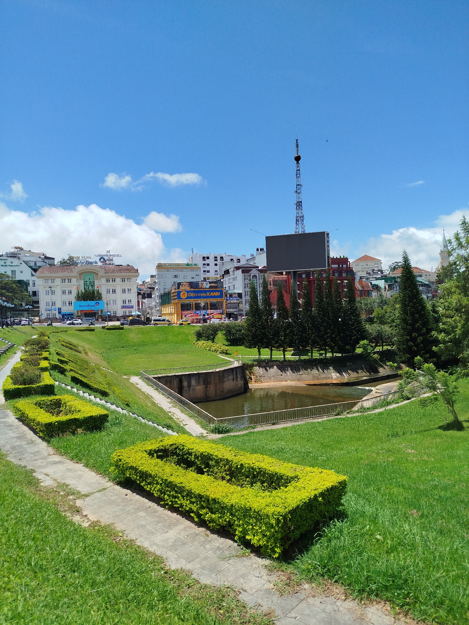 This is a daytime, eye-level, outdoor, wide-angle shot of a park in Da Lat, Vietnam. In the foreground, a paved walkway curves through bright green grass, bordered by neatly trimmed, rectangular hedges. A small, shallow canal runs alongside the walkway, with concrete retaining walls. Beyond the canal, the ground slopes upward, covered in grass and more manicured hedges.
In the middle ground, a row of tall, dark green pine trees stands behind the canal. Behind the trees, a city skyline is visible. Several buildings of varying heights and architectural styles are present, including a white building with the VietinBank logo. A large, blank, dark digital billboard is mounted on a tall metal tower in the distance. A few cars and vans are visible on the street in front of the buildings.
The sky is a clear, bright blue with scattered white cumulus clouds. The overall impression is of a sunny day in a well-maintained urban park. The area appears to be a public space, possibly a promenade or a section of a larger park adjacent to the city.