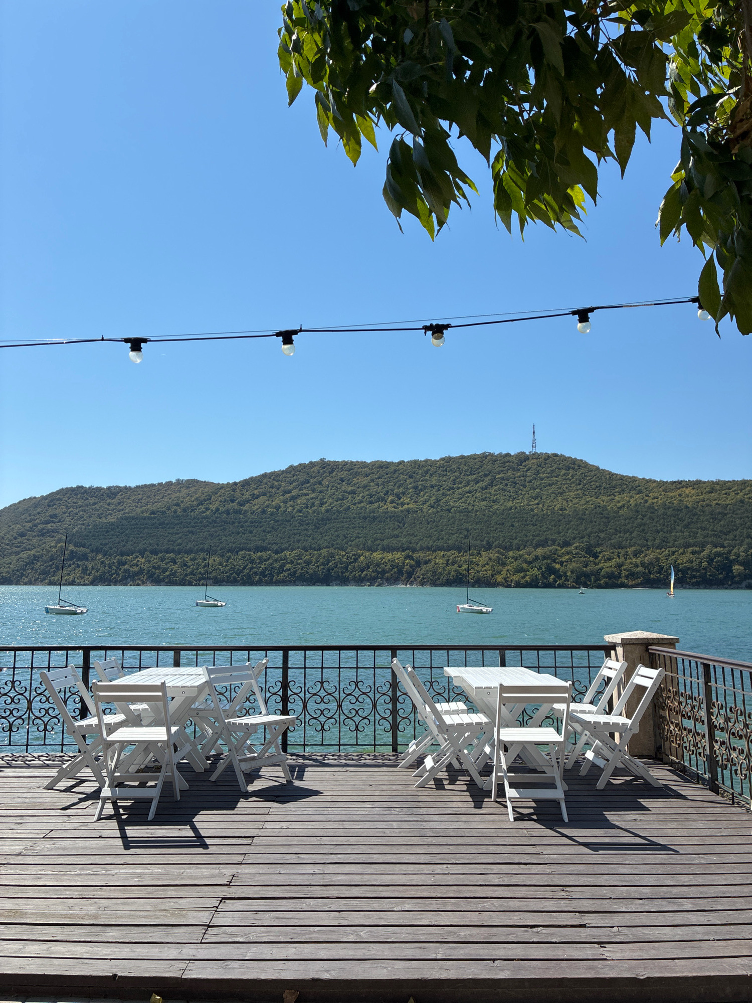The image shows an outdoor seating area with white wooden tables and chairs on a wooden deck overlooking a body of water. In the background, a green, tree-covered hill rises from the water's edge. Several sailboats are visible on the water. String lights with exposed bulbs are suspended overhead, and green leaves hang from a tree in the upper right corner. The scene appears to be a cafe or restaurant terrace in Abrau-Dyurso, Russia, likely overlooking Abrau Lake. The sky is clear and blue. There are no people visible in the image.